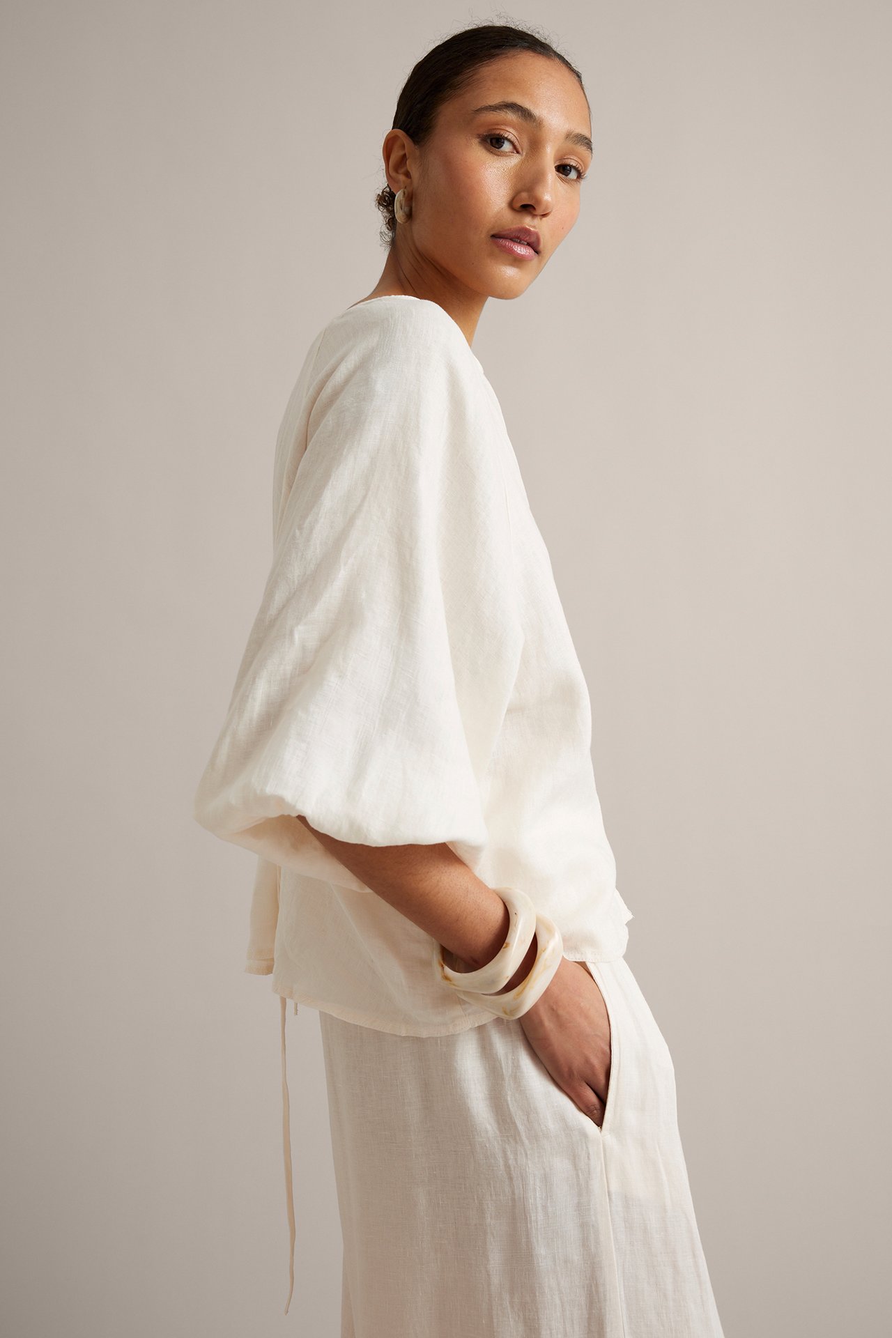 Bok widok kobiety w białej lnianej bluzce z szerokimi rękawami, wiązaniami, lnianymi spodniami, bransoletkami i kolczykami.