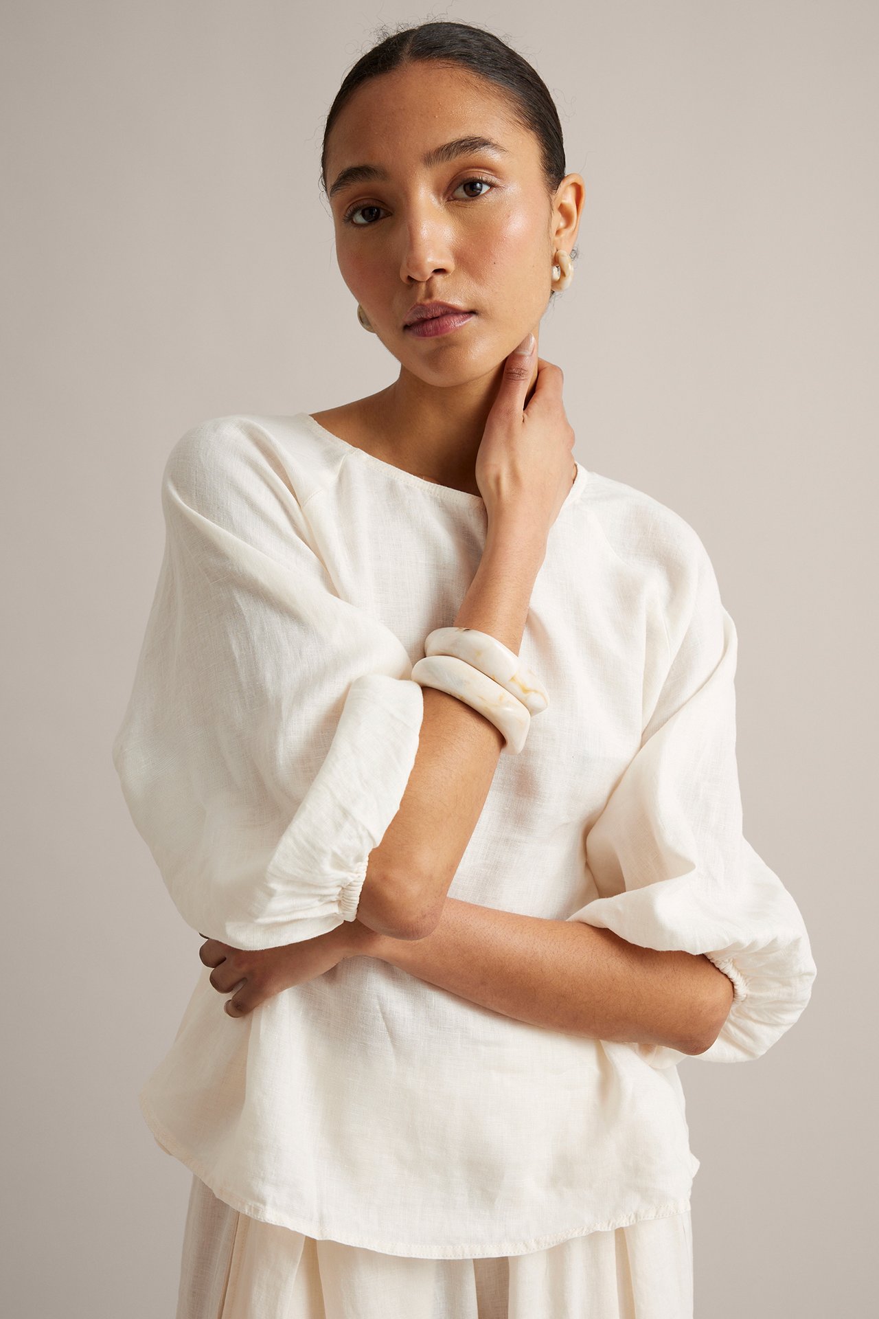 Widok z przodu: Kobieta w białej lnianej bluzce z szerokimi rękawami, jasnej spódnicy, kolczykach i bransoletkach.