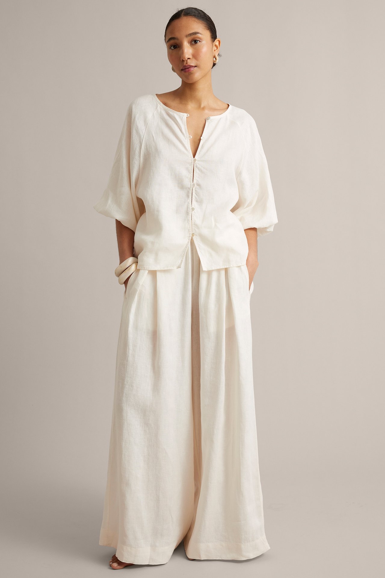 Widok z przodu: Kobieta nosi luźną białą lnianą bluzkę, szerokie lniane spodnie, białe bransoletki, złote kolczyki.