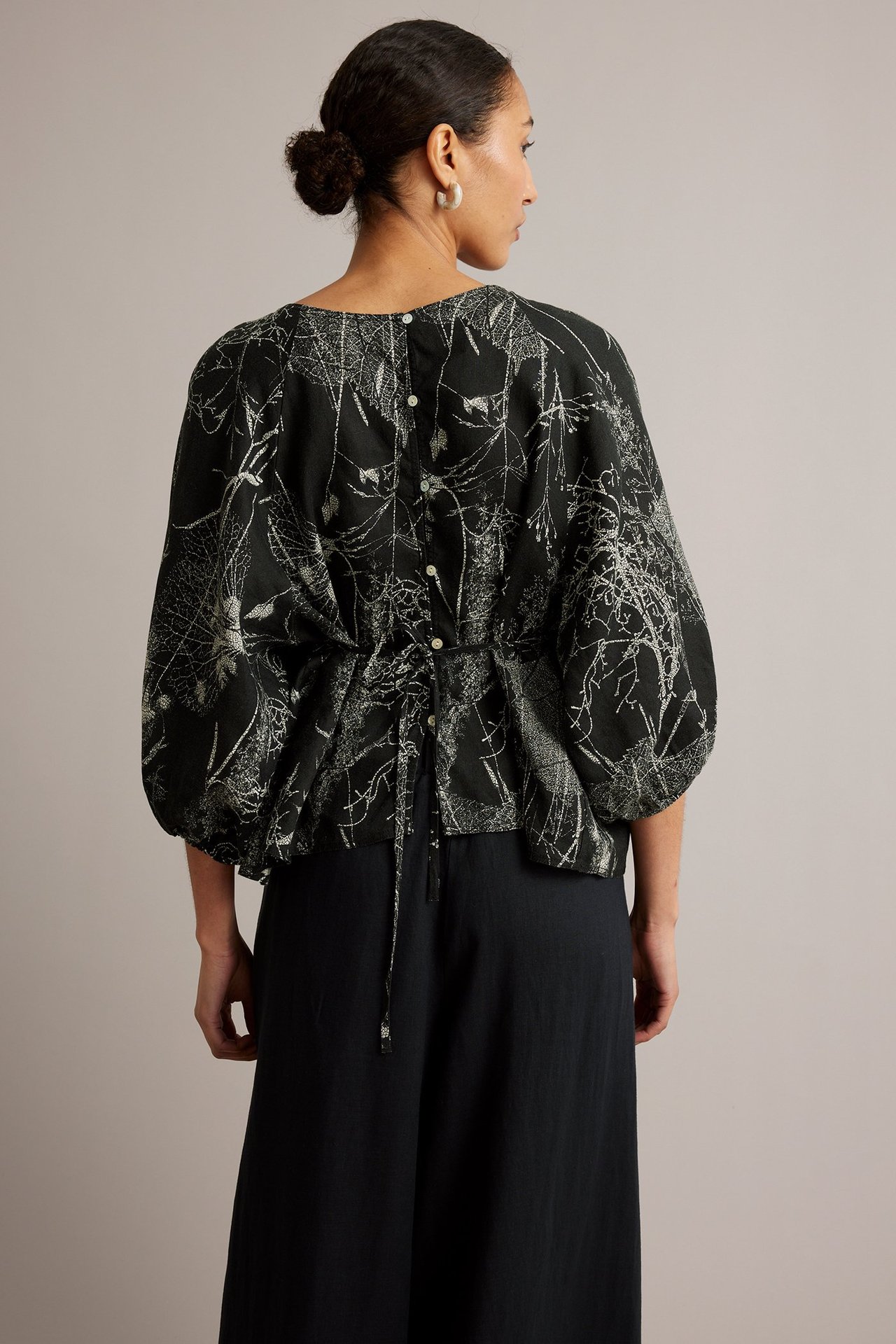 Widok z tyłu: kobieta w czarnej lnianej bluzce w kwiaty, z wiązaniem, szerokimi rękawami i czarnymi spodniami.