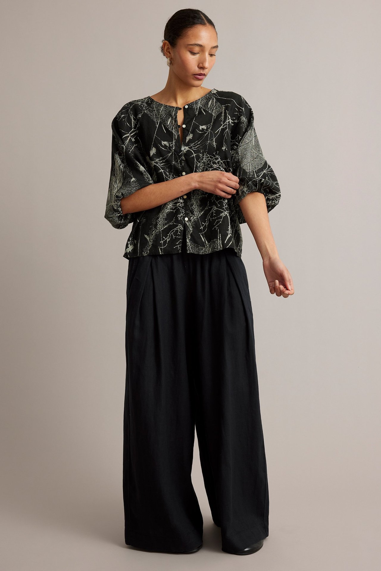 Edestäpäin kuvattu nainen mustassa pellavapuserossa, jossa vaalea kukkakuvio, ja mustissa leveälahkeisissa housuissa.