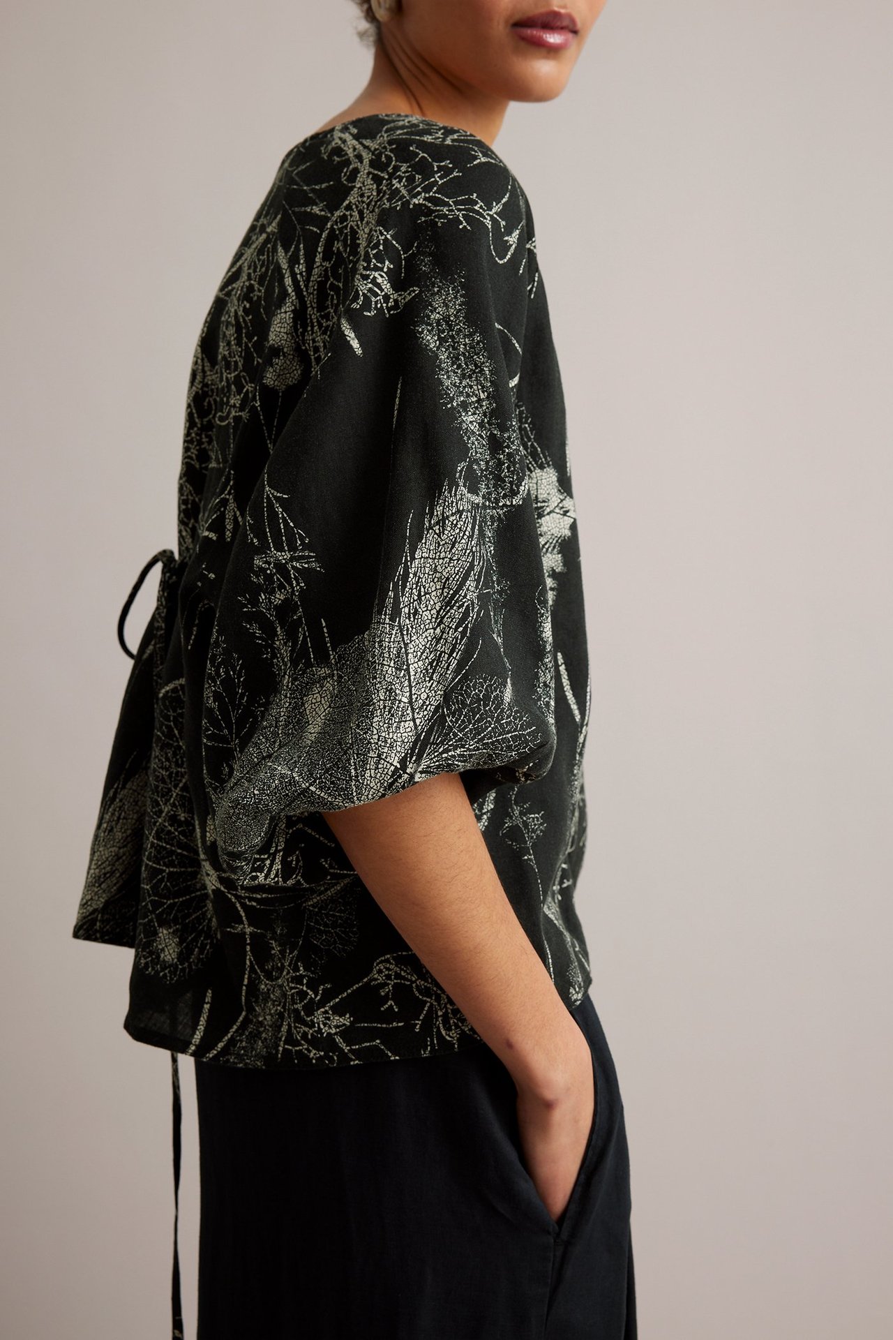 Widok z boku kobiety w czarnej lnianej bluzce z jasnym kwiatowym wzorem i wiązaniem, do tego czarne spodnie.