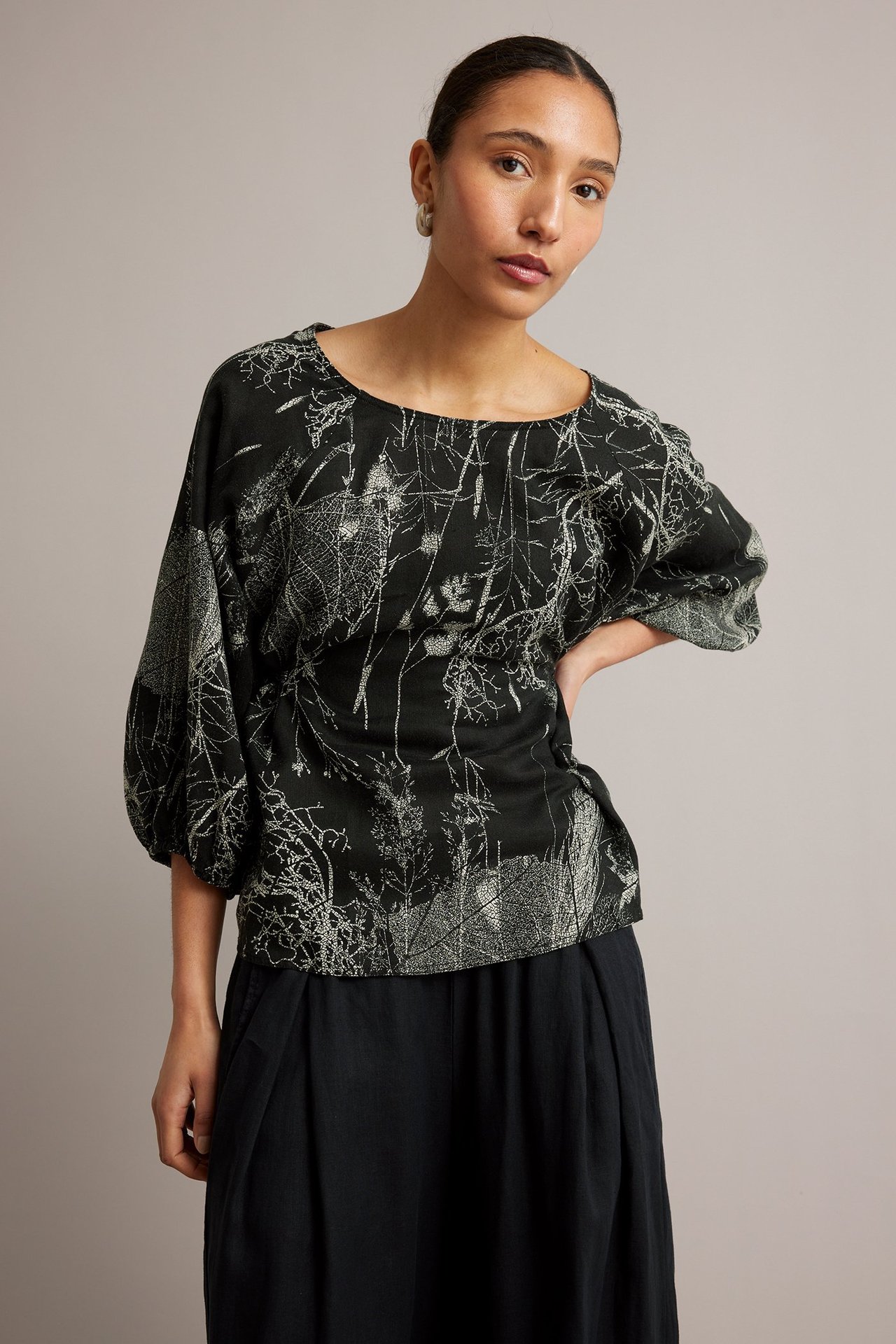 Z przodu: kobieta w czarnej lnianej bluzce z jasnoszarym botanicznym wzorem i rękawami, w czarnych spodniach.