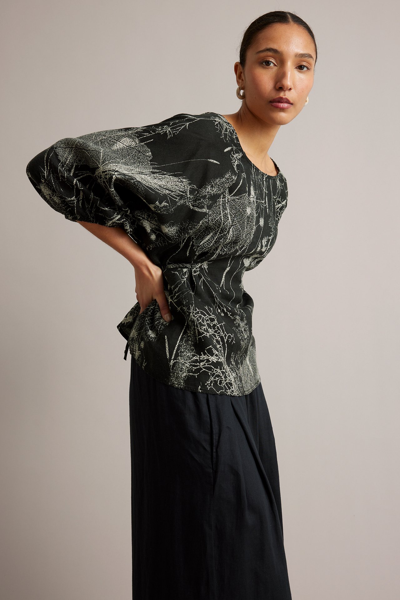 Widok z boku kobiety w czarnej lnianej bluzce z jasnym botanicznym wzorem i wiązaniem, z czarnymi spodniami.