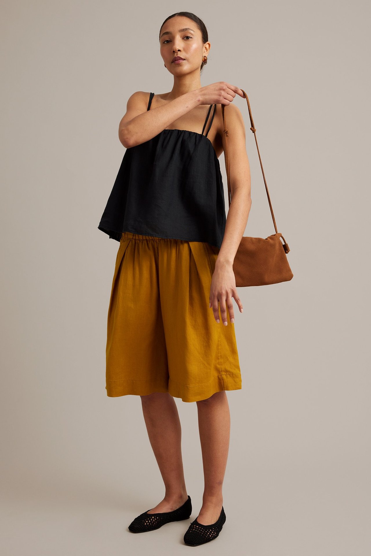 Framifrån: Kvinna i gula plisserade linneshorts, svart linne, brun axelremsväska och svarta ballerinaskor.