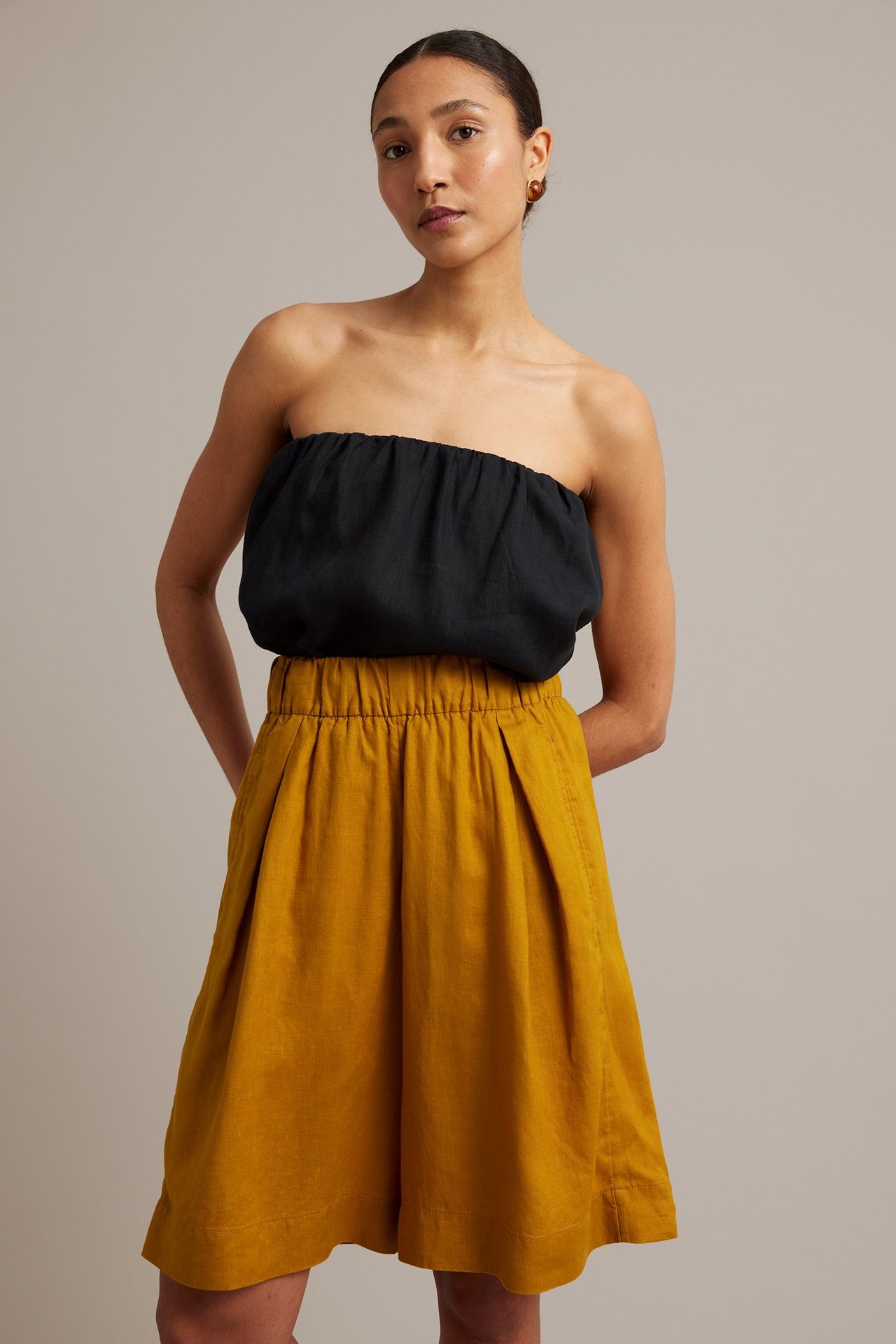 Front view: Kvinna bär gula knälånga linneshorts med resår i midjan, sidfickor och en svart bandeau-topp.