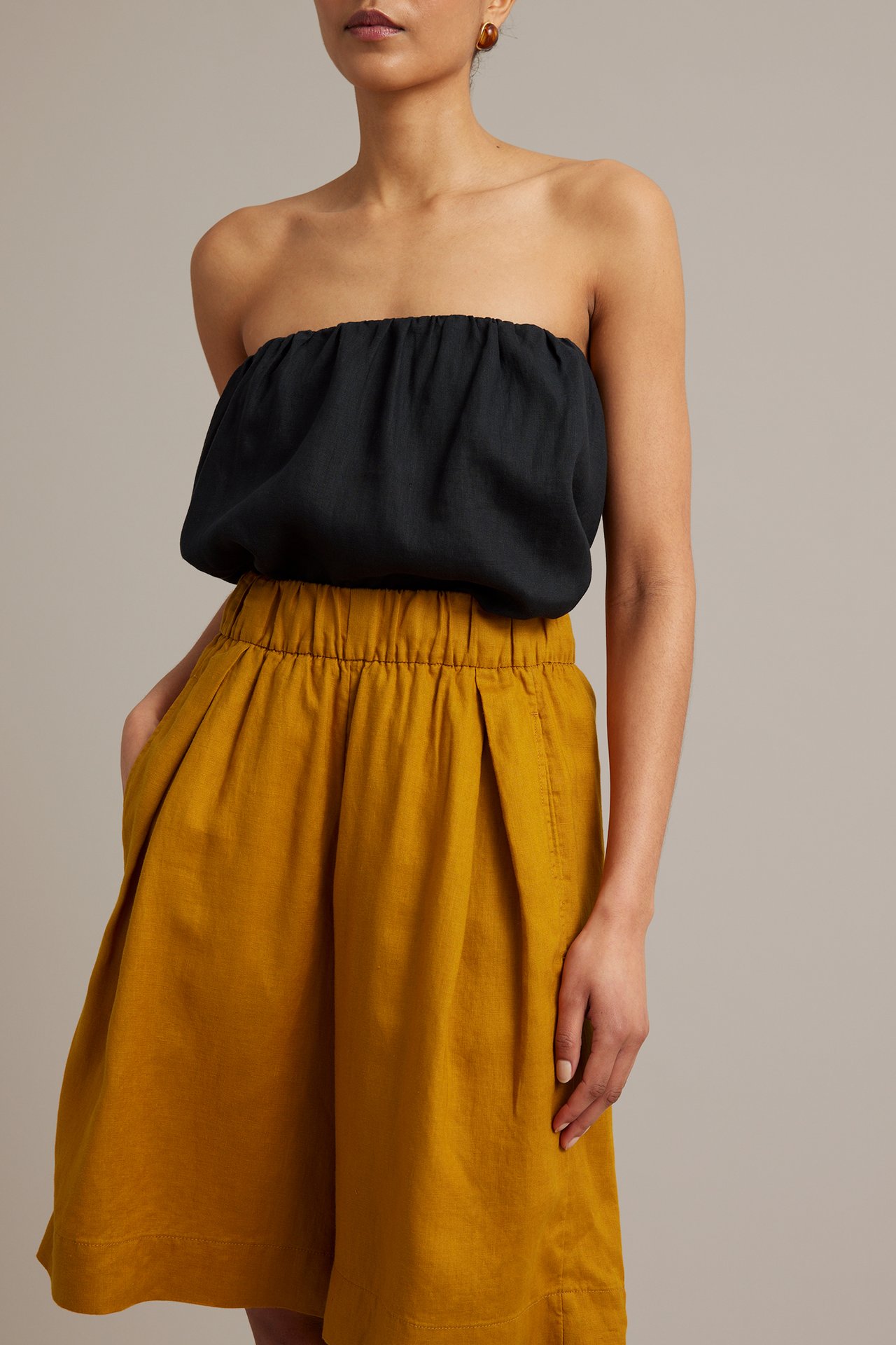 Framifrån: Kvinna i gula linnebermudashorts med resår i midjan och sidfickor, med svart bandeau-topp.