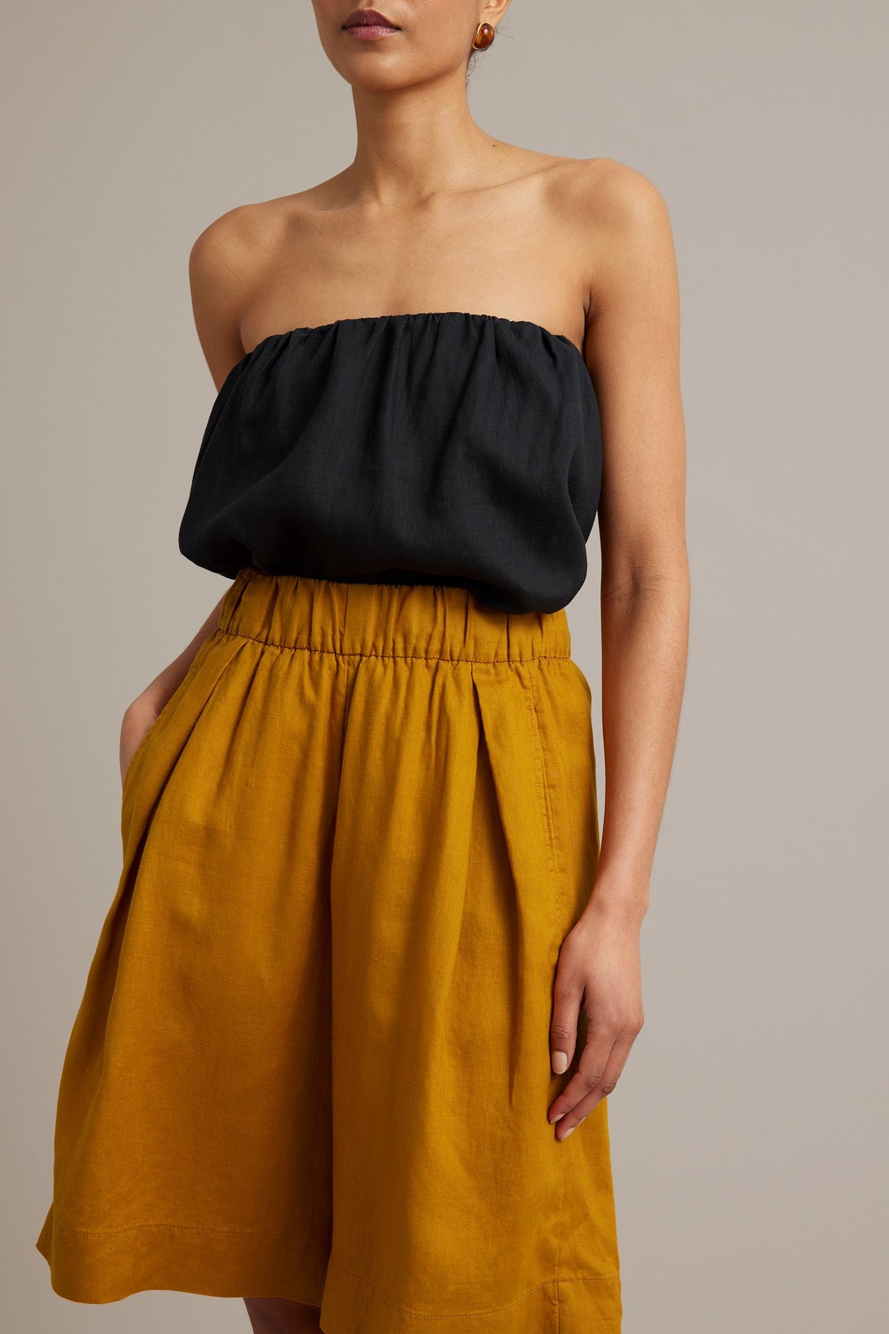 Framifrån: Kvinna i gula linnebermudashorts med resår i midjan och sidfickor, med svart bandeau-topp.