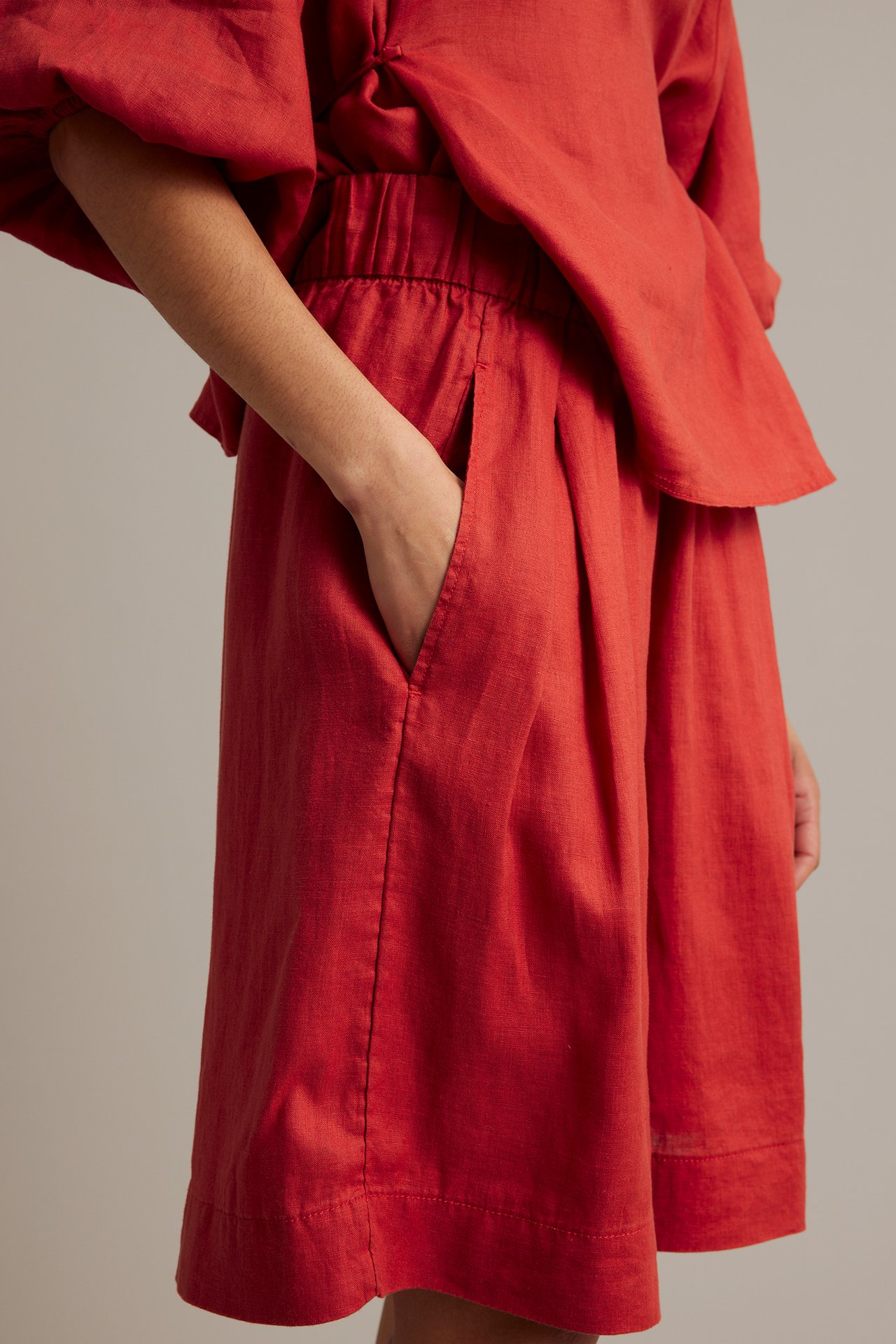 Sidovy: Röda knälånga linneshorts med resår i midjan, kvinnas hand i fickan, burna med matchande topp.