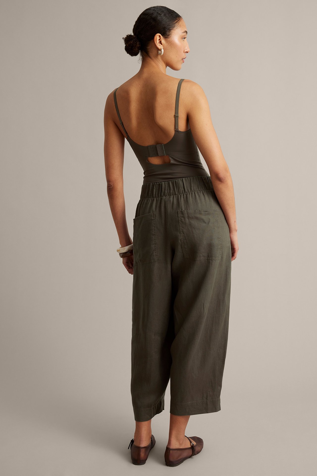 Kvinna, bakifrån, i mörkgröna barrel-fit linnebyxor, matchande topp, vitt armband och bruna skor.