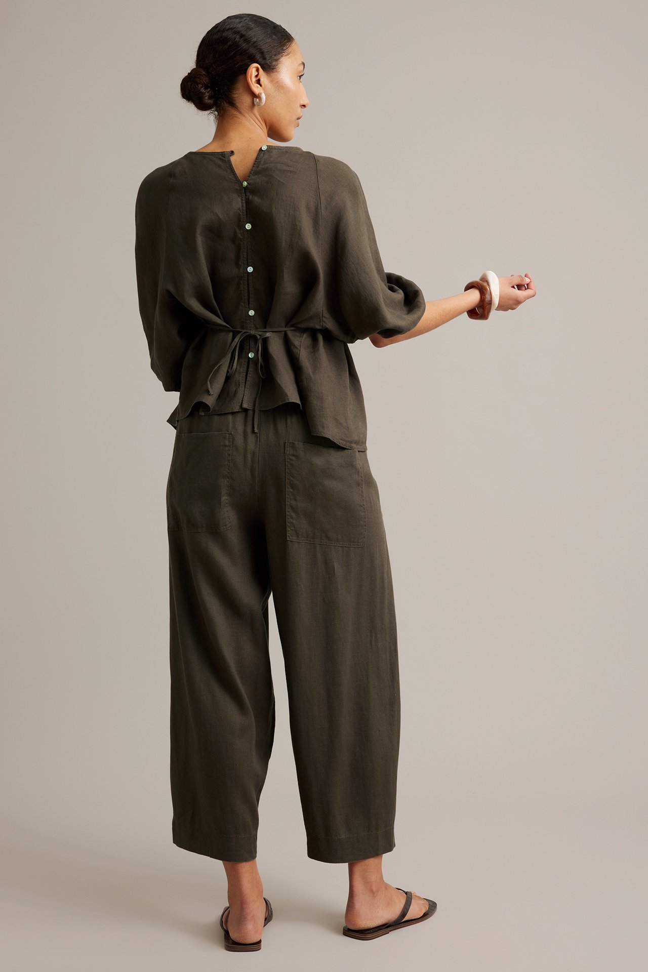 Naisen takakuva ruskeissa barrel-fit pellavahousuissa, yhteensopivassa puserossa ja sandaaleissa.