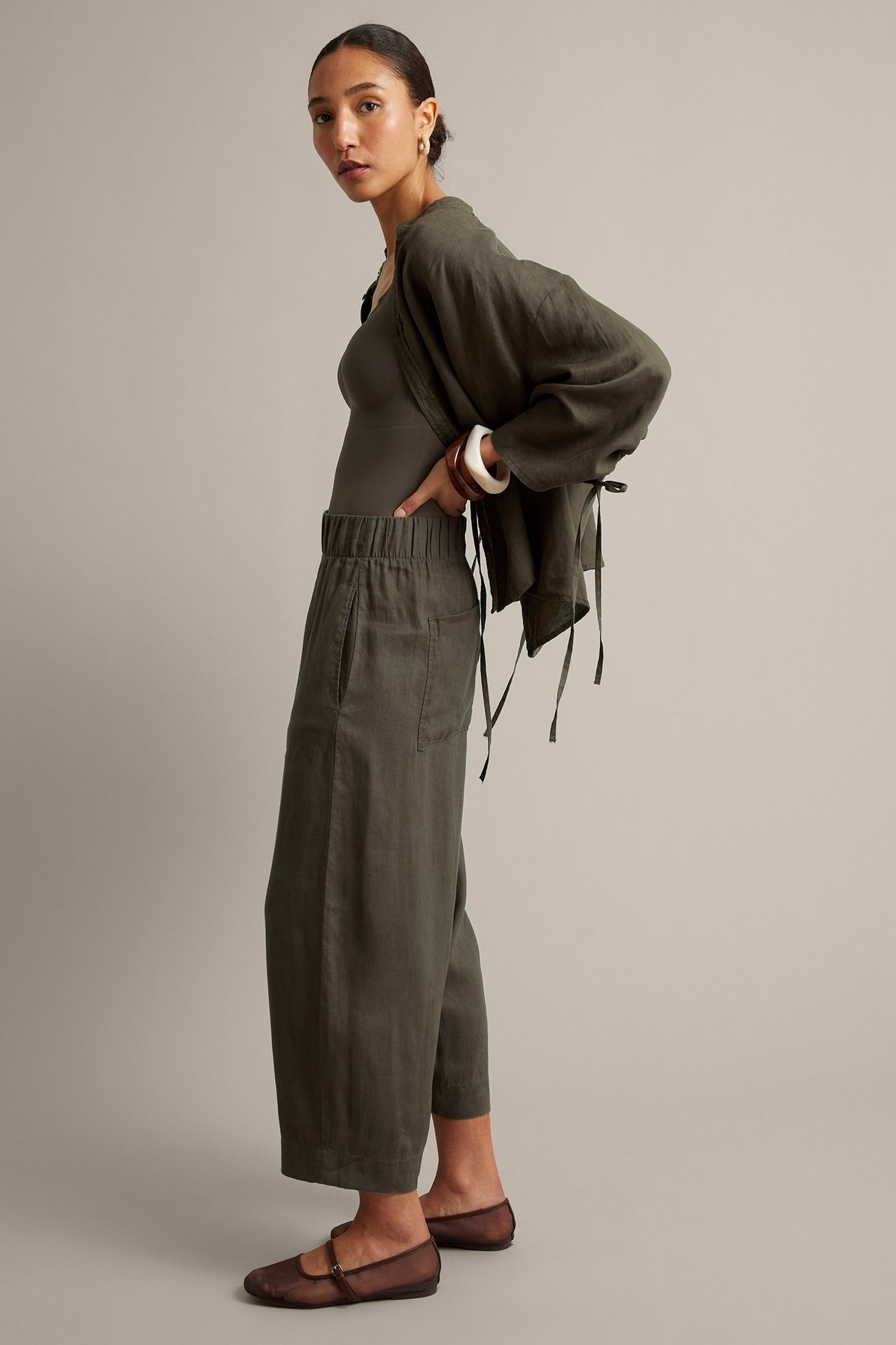 Widok z boku: Kobieta w brązowych lnianych spodniach barrel fit z elastycznym pasem, ciemnym topie i otwartej kurtce.