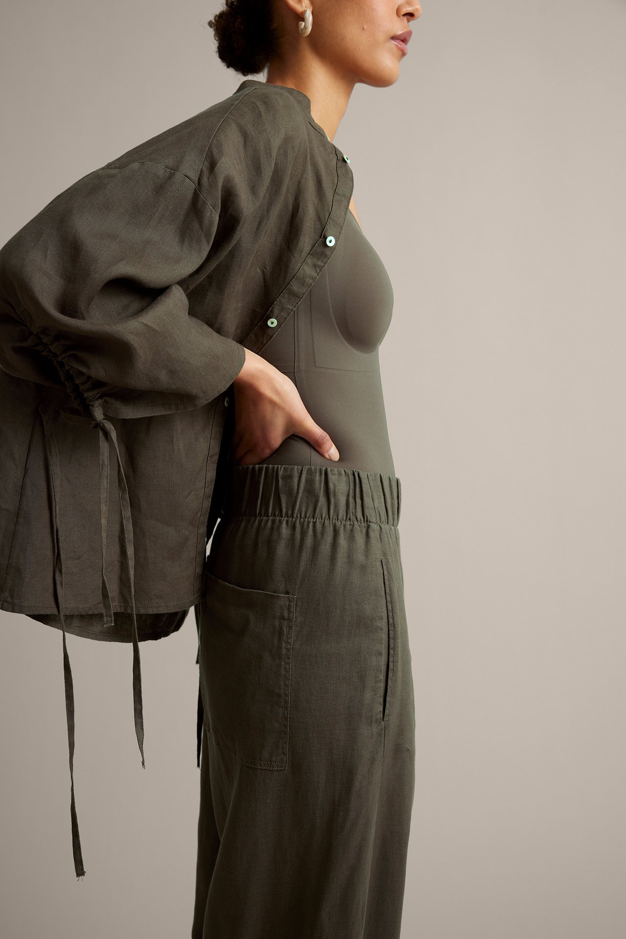 Kvinna i olivgröna linnebyxor med barrel-fit och resår i midjan, matchande öppen skjorta och olivgrön bodysuit.
