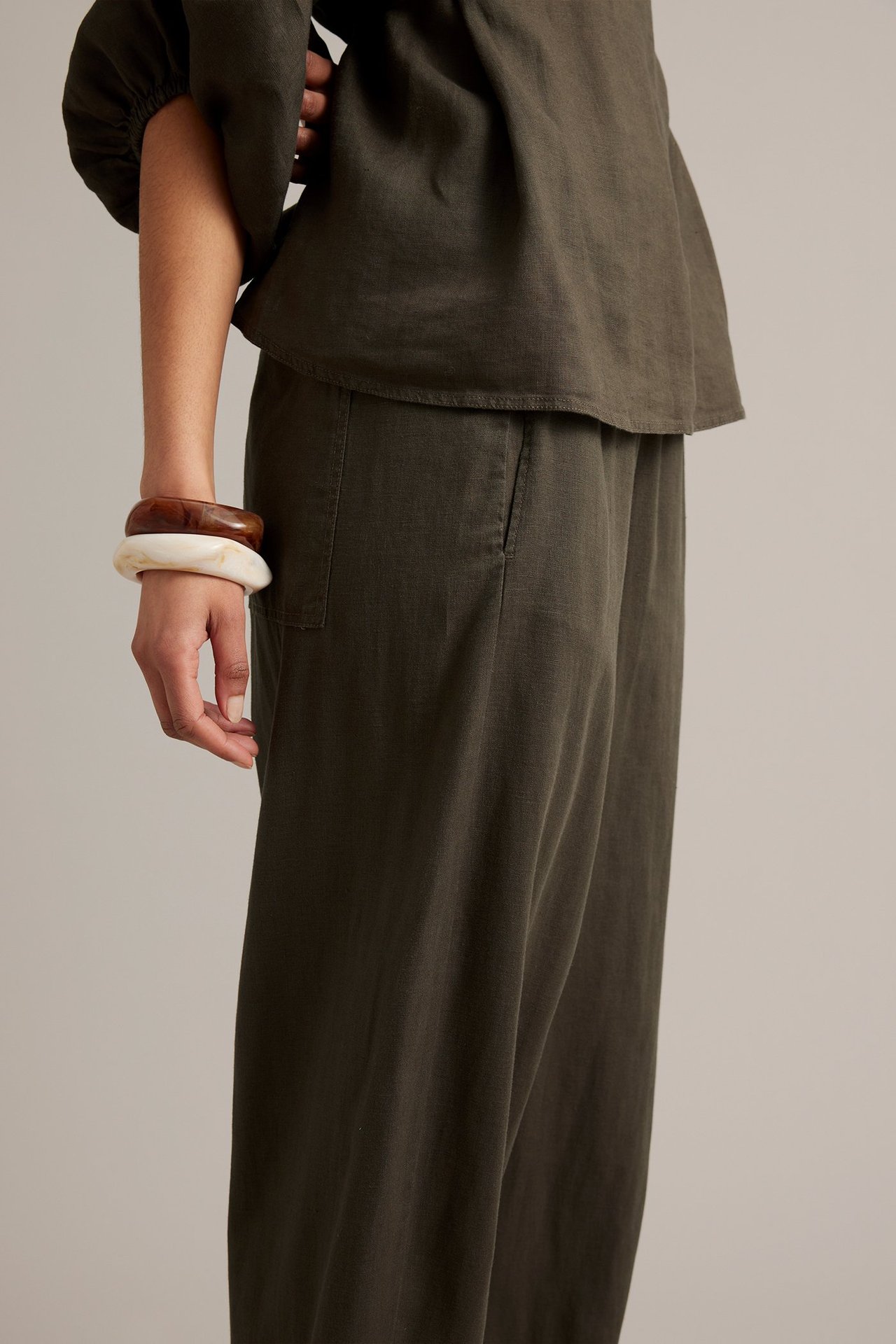 Nainen sivulta, yllään ruskeat pellavahousut barrel leg -malliset, toppi ja kaksi isoa rannekorua.