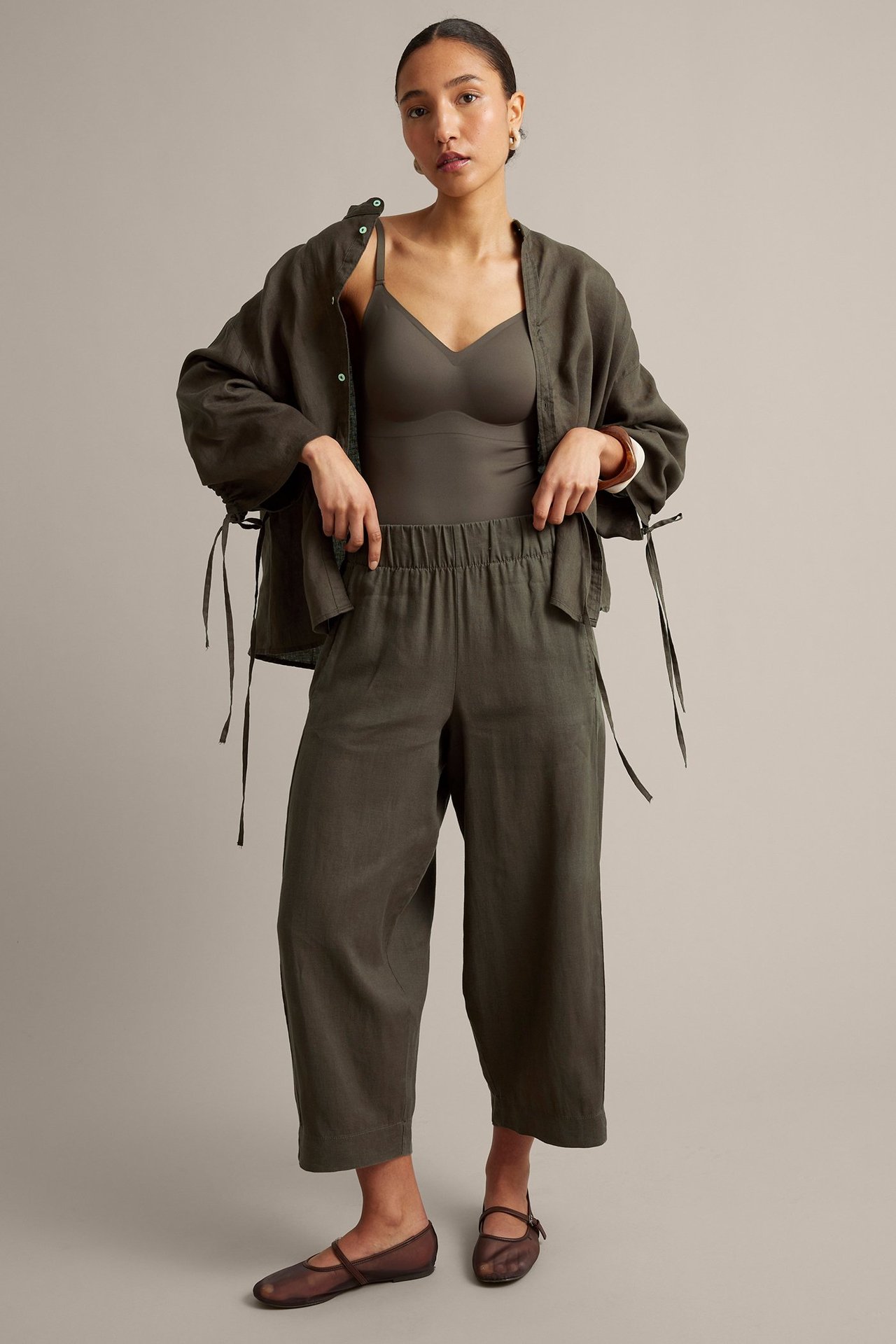Widok z przodu kobiety w ciemnooliwkowych lnianych spodniach barrel-fit, pasującej kurtce, topie i brązowych baletkach.