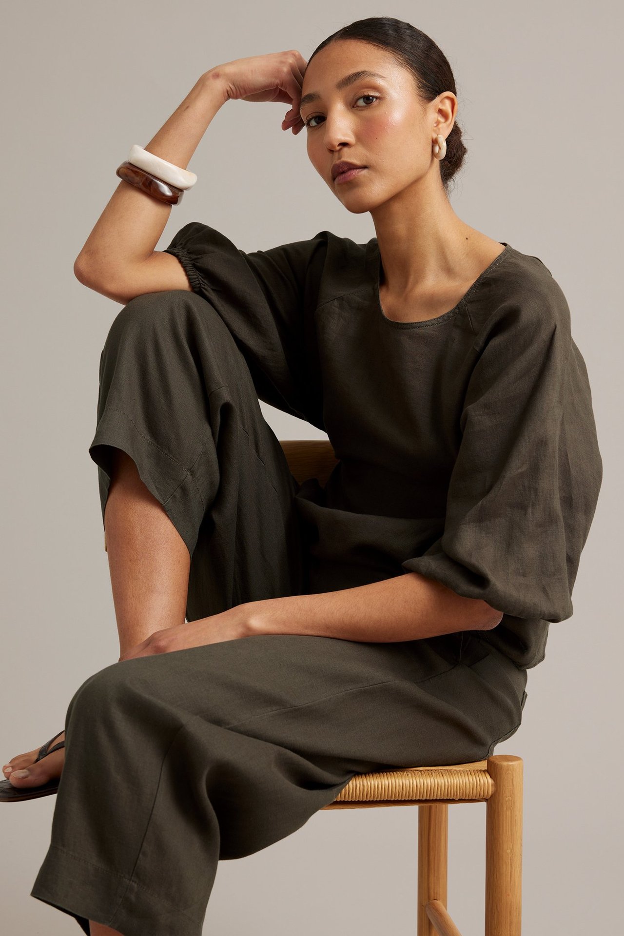 Edestä päin nainen tummanruskeissa pellavahousuissa ja yhteensopivassa topissa, rannekoruissa ja korvakoruissa.