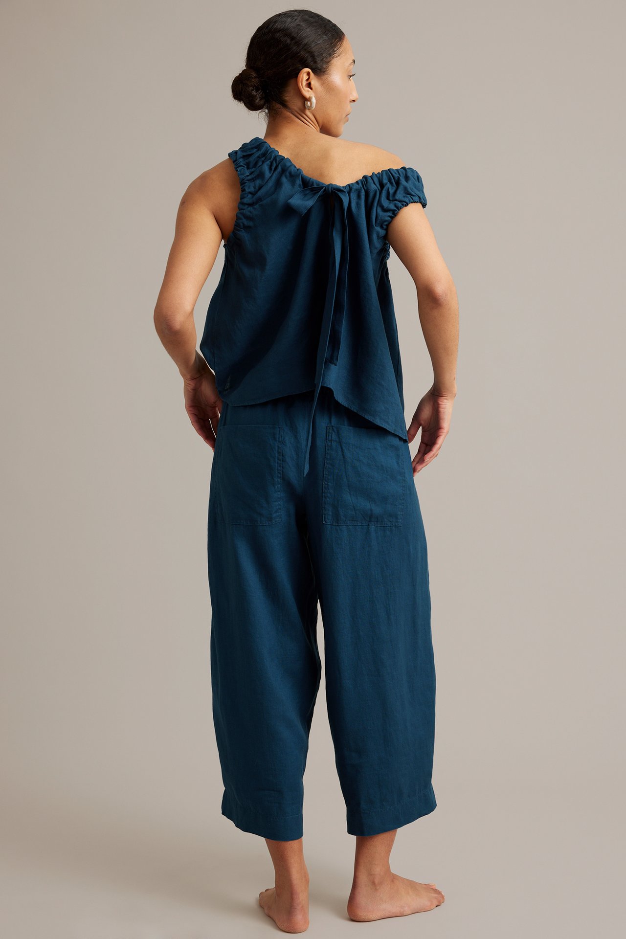 Widok z tyłu kobiety w niebieskich lnianych spodniach barrel fit z tylnymi kieszeniami i topie off-shoulder.