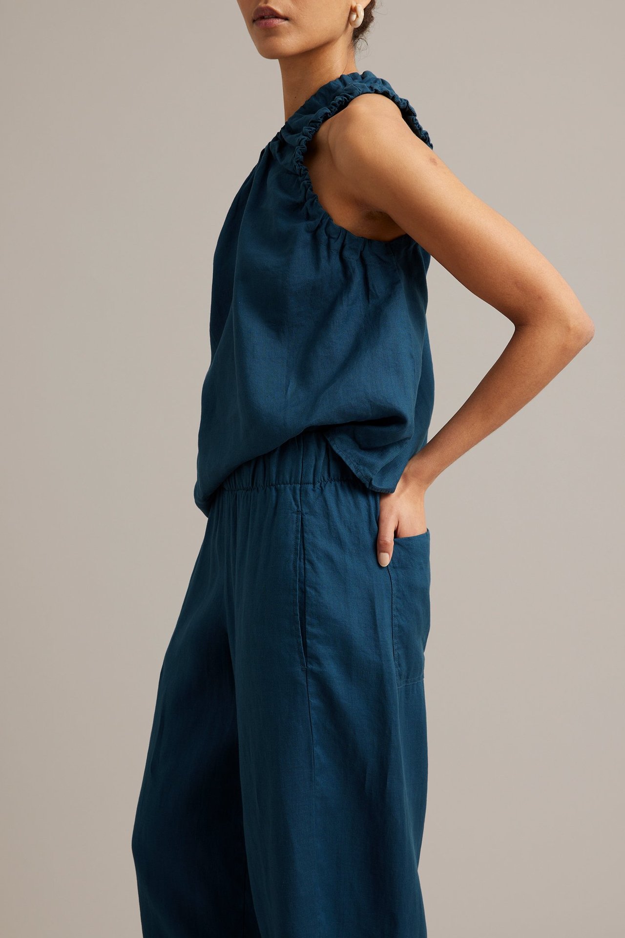 Naisen sivukuva sinisissä pellavahousuissa, joissa barrel-lahkeet, kuminauhavyötärö ja yhteensopiva hihaton toppi.