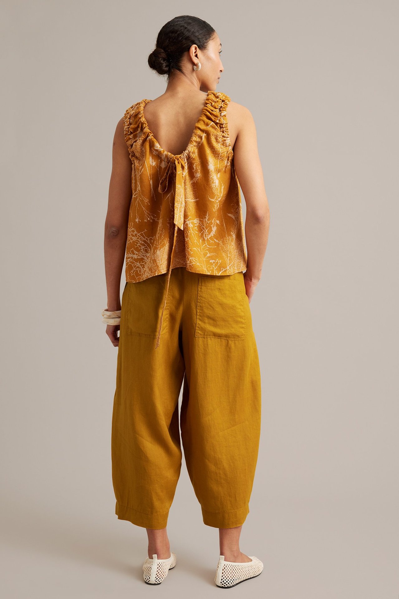 Bakifrån: kvinna i gula linnebyxor med barrel fit och gul mönstrad ärmlös topp med knytband bak.