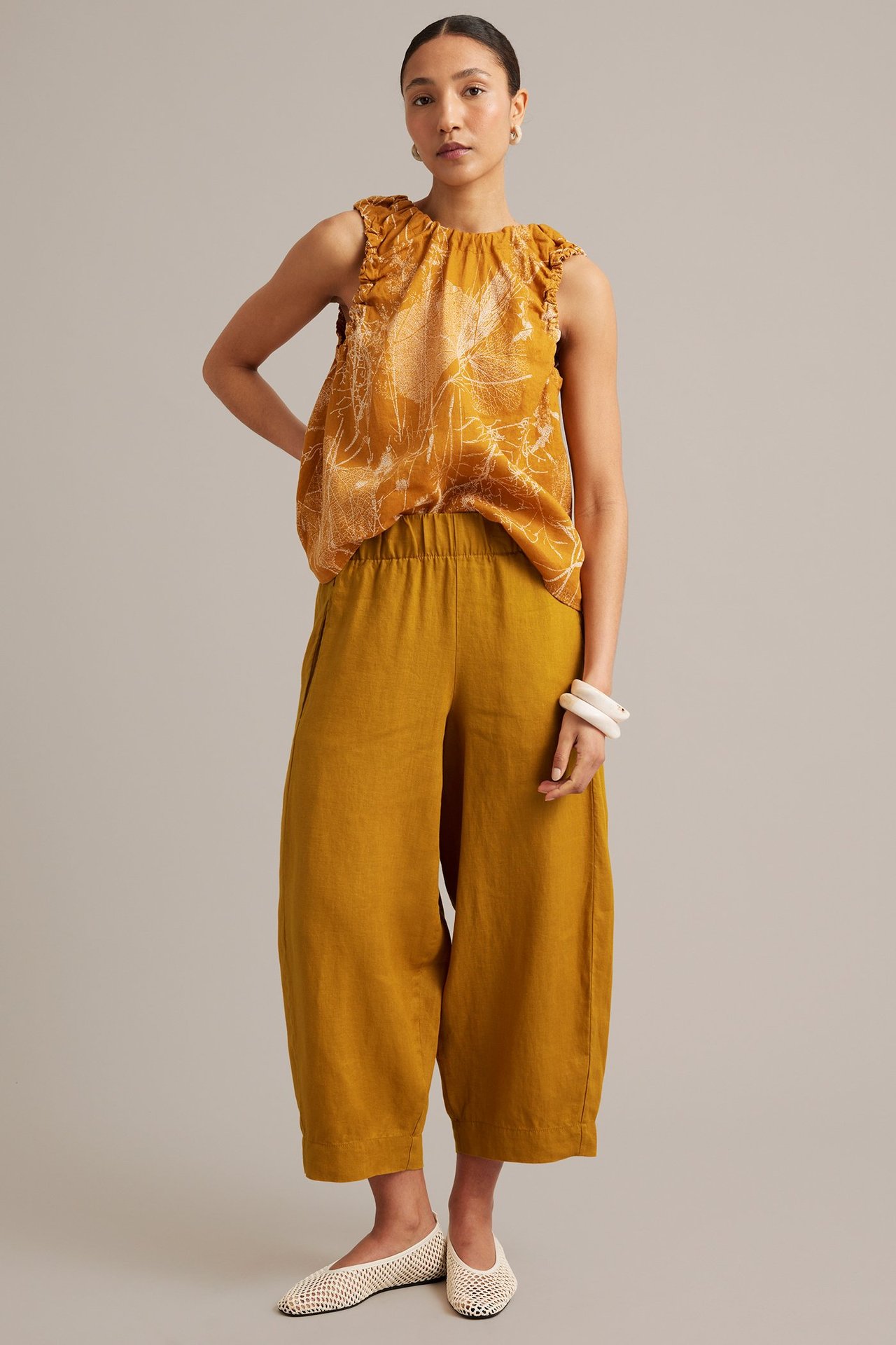 Widok z przodu: Kobieta w żółtych lnianych spodniach barrel fit, wzorzystym topie i białych siateczkowych balerinach.