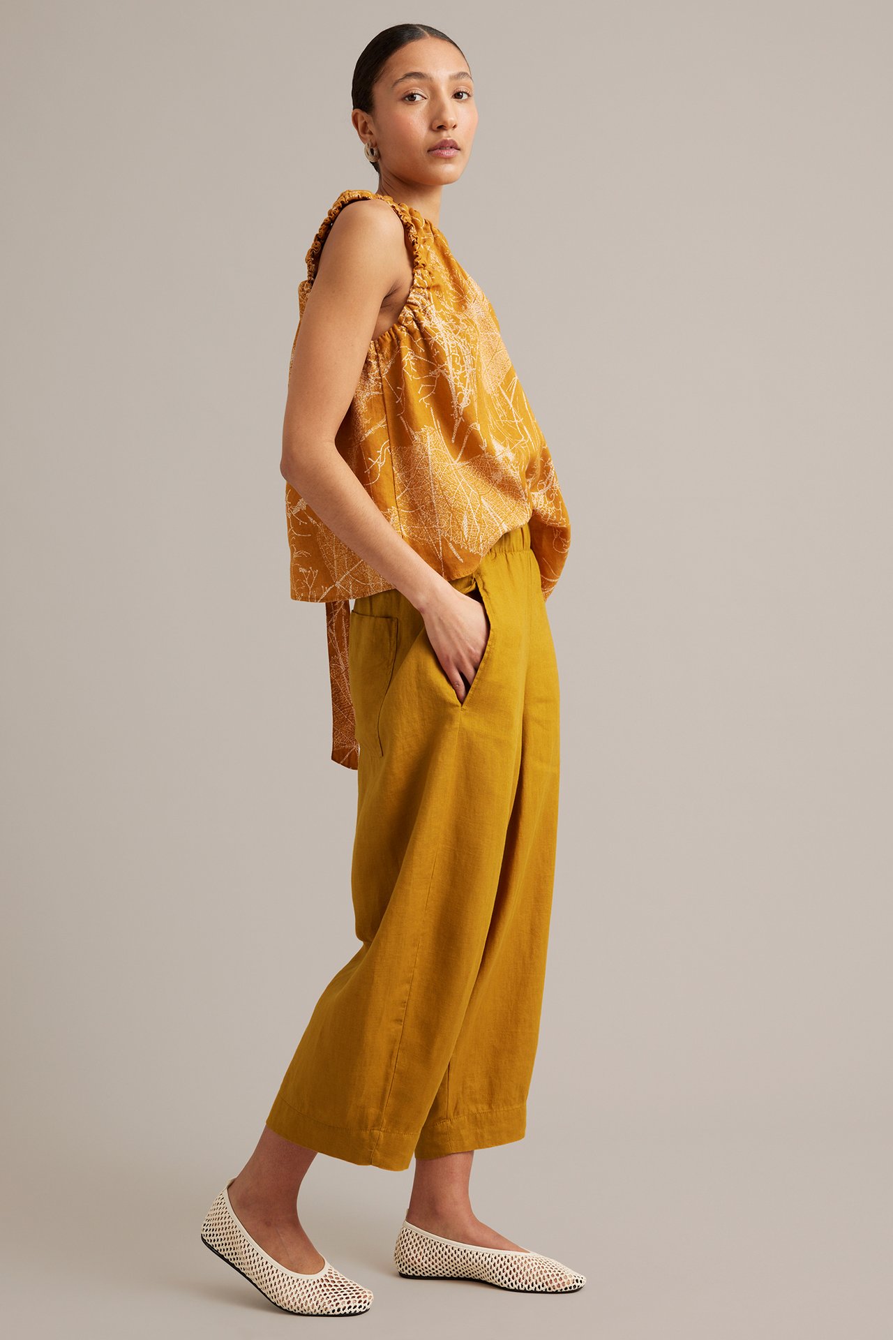 Sidovy av kvinna i senapsgula barrel-leg linnebyxor, mönstrad ärmlös topp och ljusa nätballerinor.
