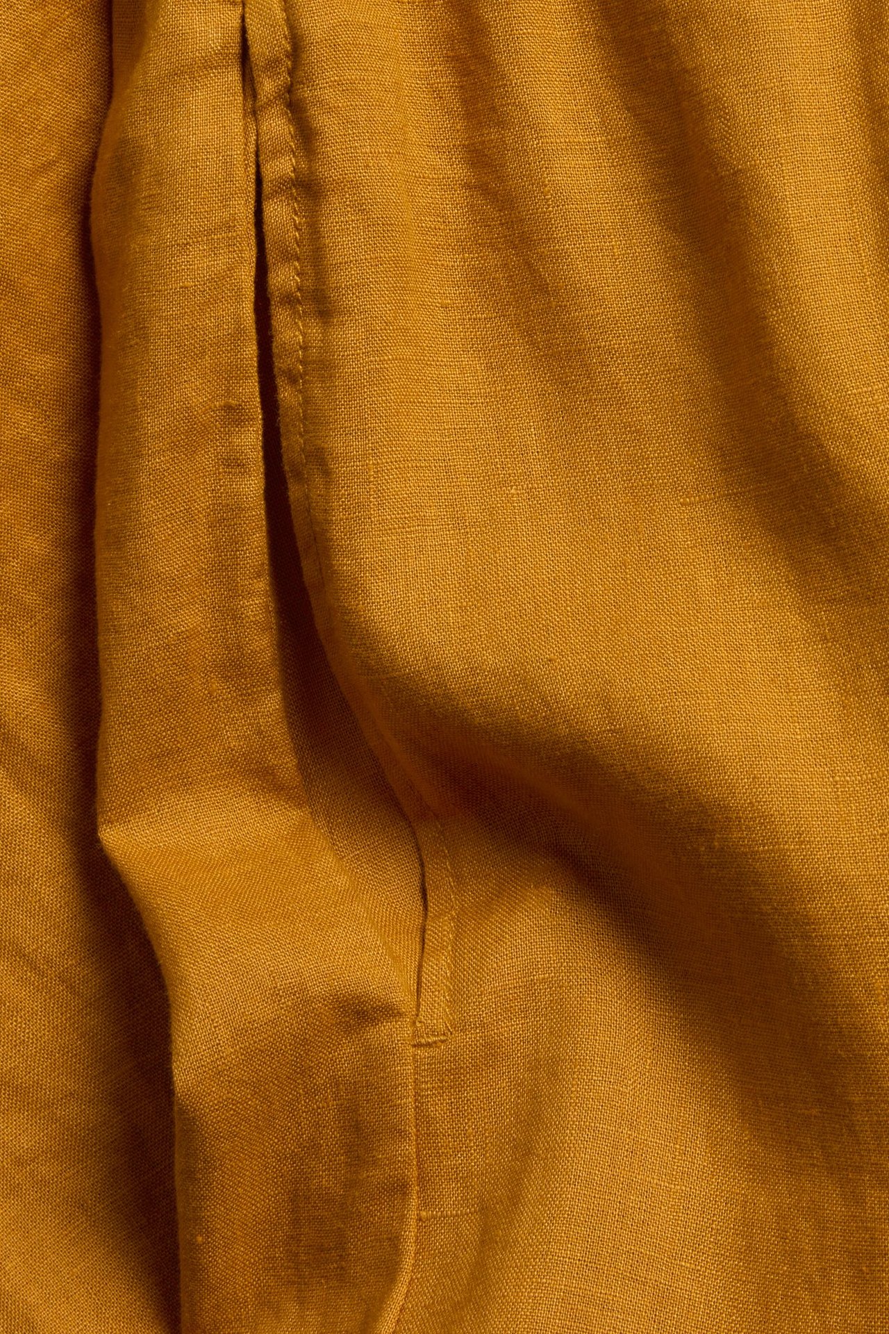 Zbliżenie musztardowo-żółtej tkaniny lnianej spodni, ukazujące jej teksturę i szwy.