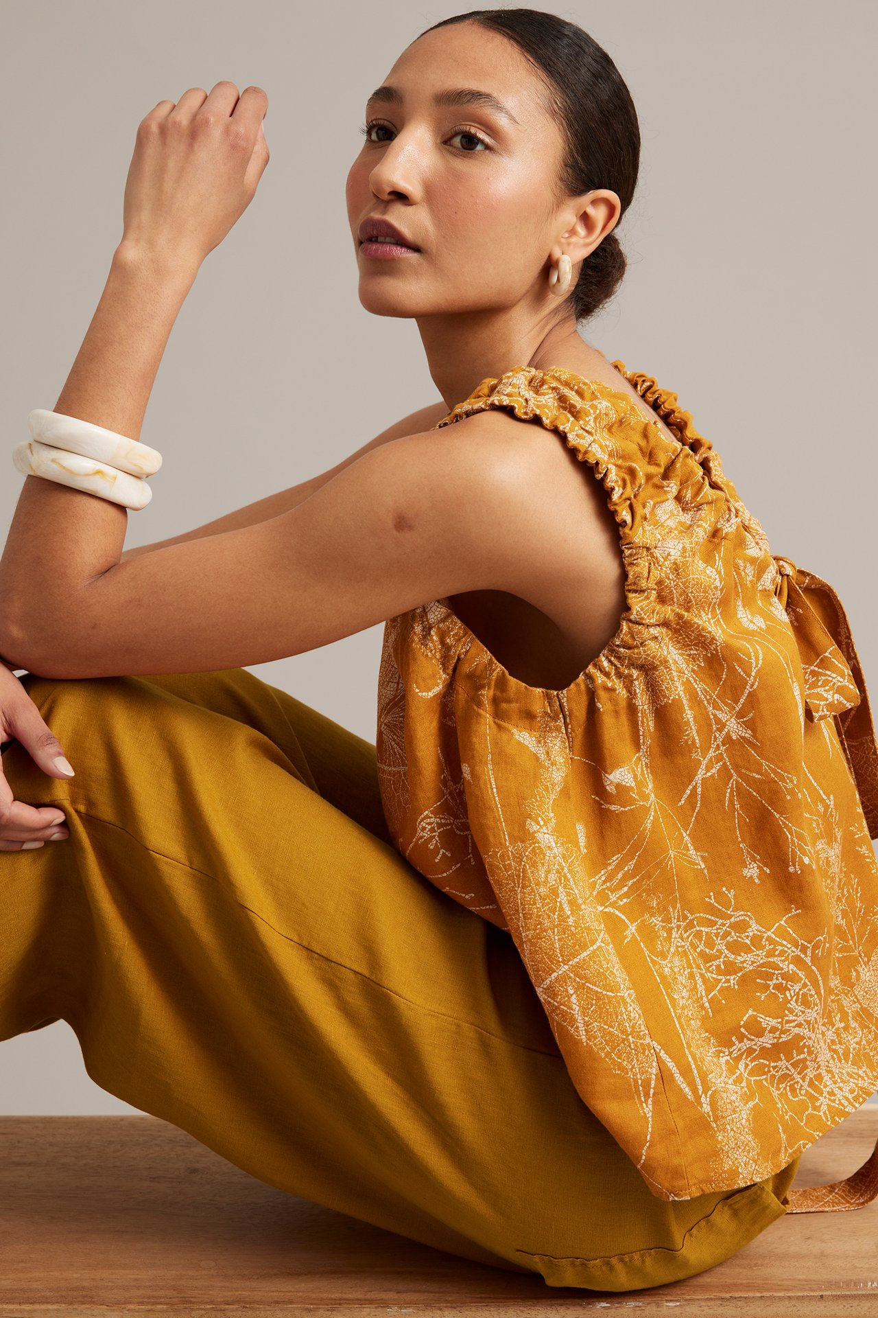 Sidovy: kvinna i gula linnebyxor med barrel fit, mönstrad gul topp, krämfärgade bangles, ringörhängen.