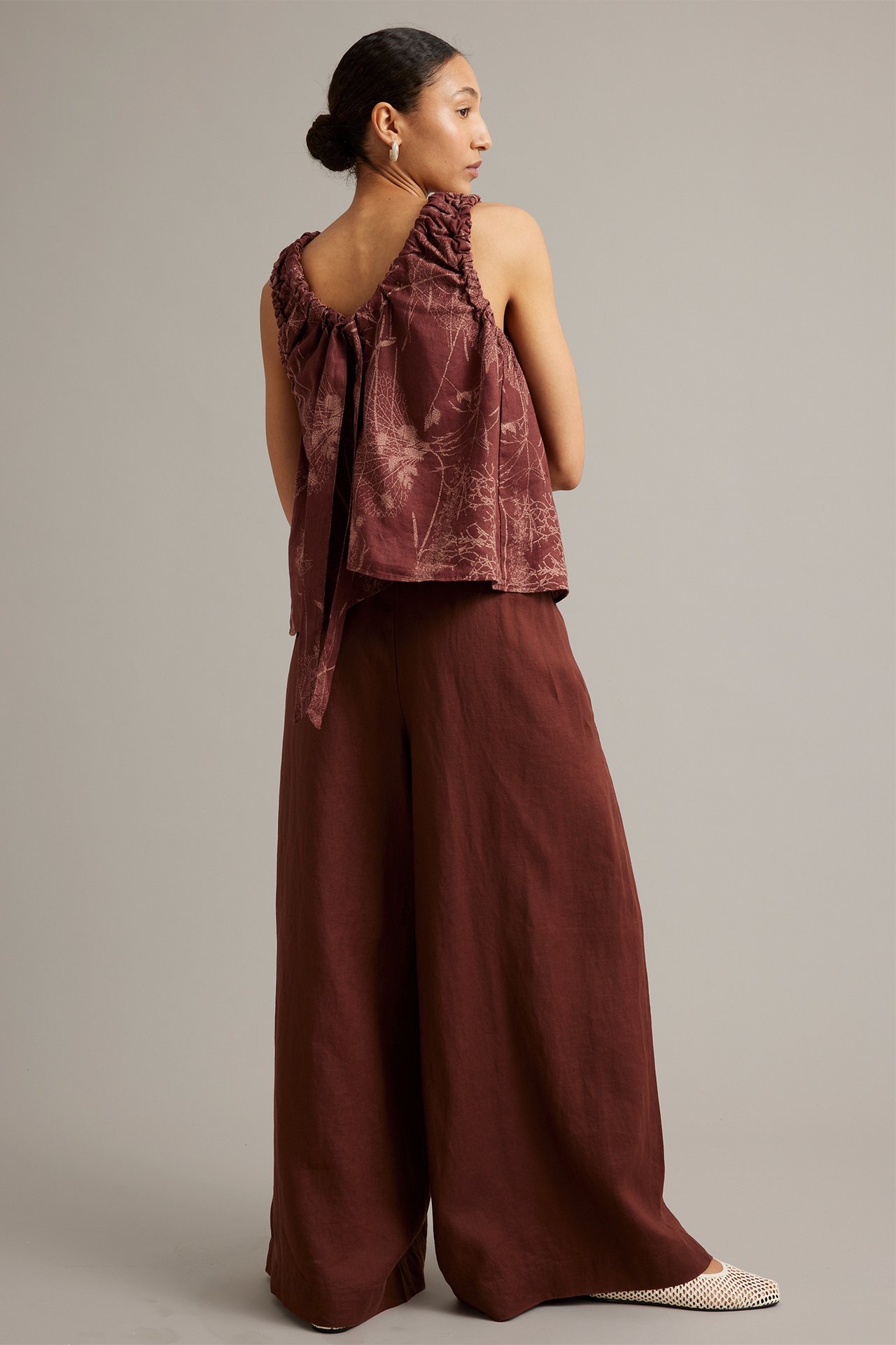 Vinklet bakfra-bilde av kvinne i vide vinrøde linbukser, mønstret brun topp og hvite vevde sko.