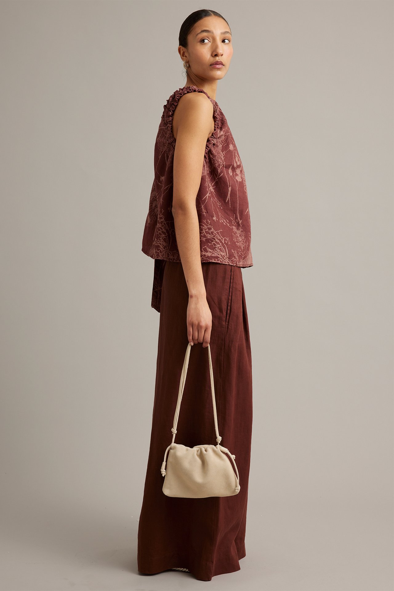 Sidebilde av kvinne i vinrød vid linbukse, mønstret ermeløs topp, holder en beige håndveske.