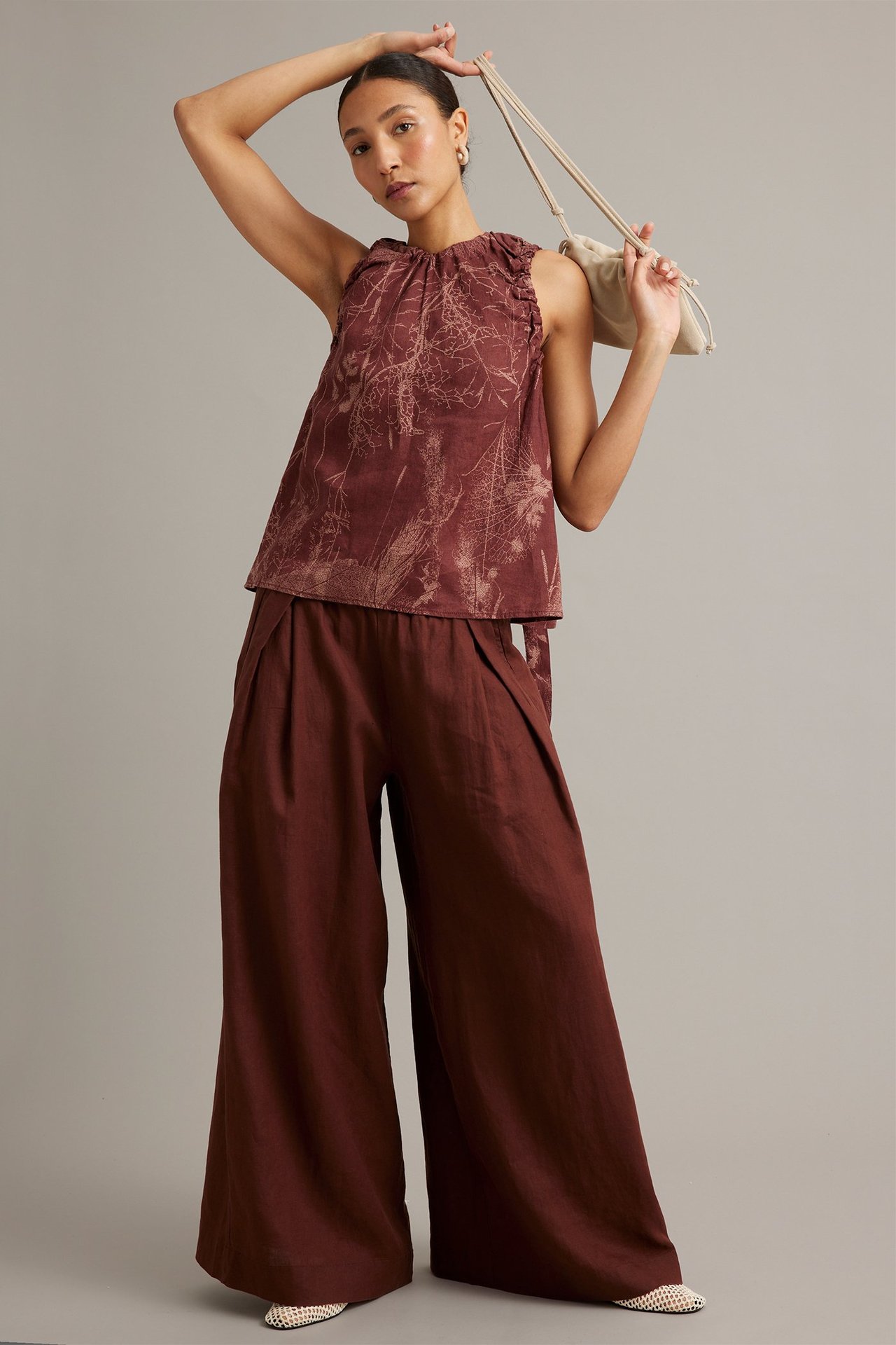 Edestä: Nainen yllään leveät viininpunaiset pellavahousut poimuilla, kuviollinen hihaton toppi ja kermanvärinen laukku.