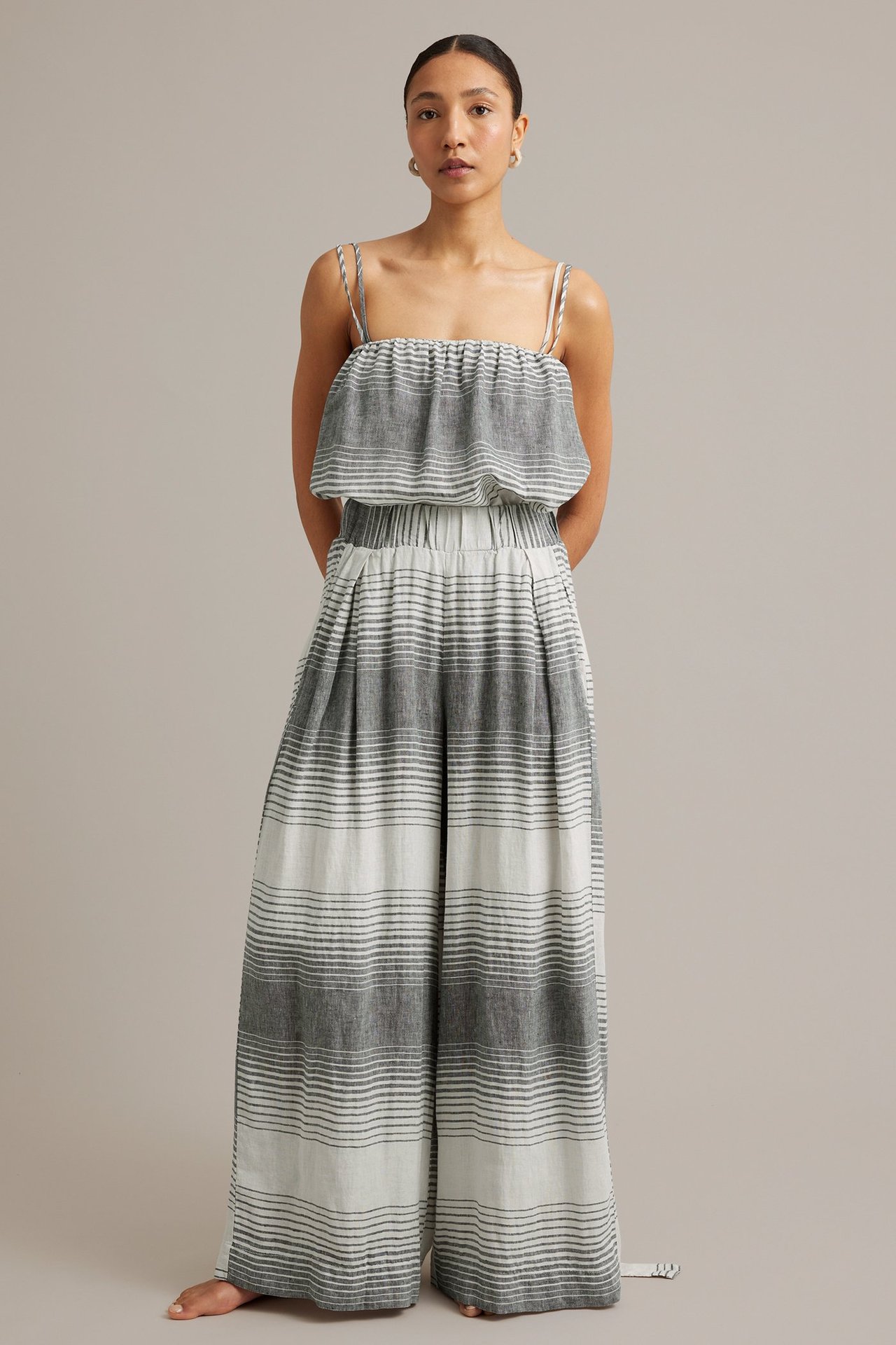 Framifrån: kvinna i grå- och vitrandiga vida linnebyxor med matchande topp med smala axelband.