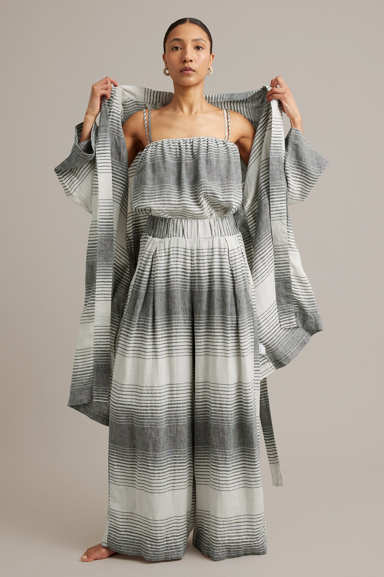 Kvinna bär mörkgrå/vitrandiga linnebyxor med vida ben, matchande topp och öppen kimono. Frontvy.