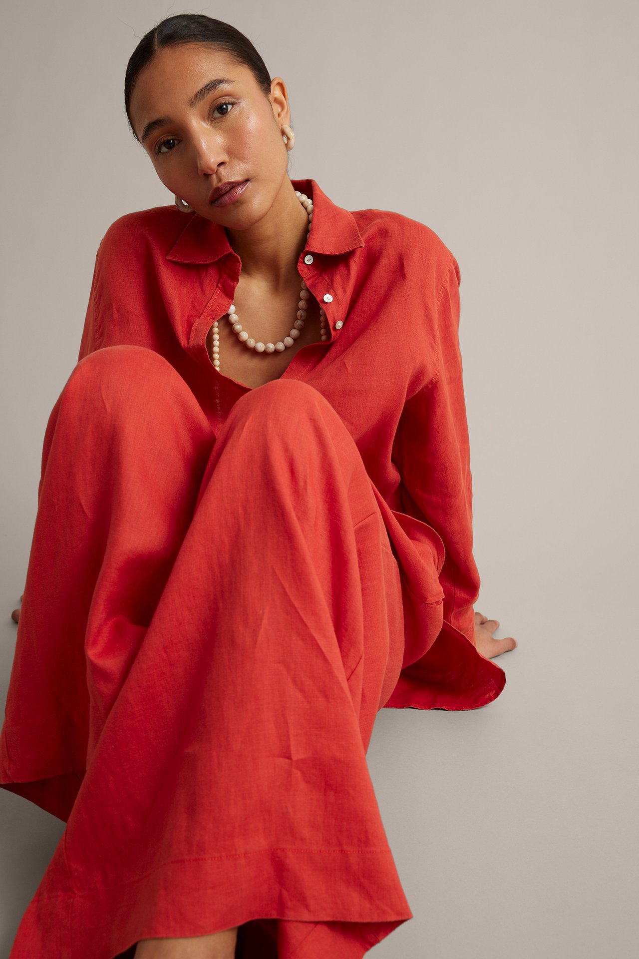 Frontalansikt: kvinne i rød vid linbukse, rød linskjorte, perlekjede og øreringer.