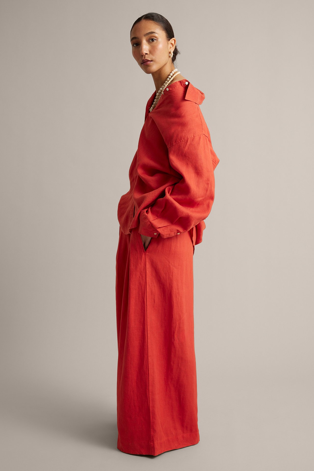 Sidebilde av kvinne i røde vide linbukser, matchende oversized linskjorte, perlekjede og øreringer.