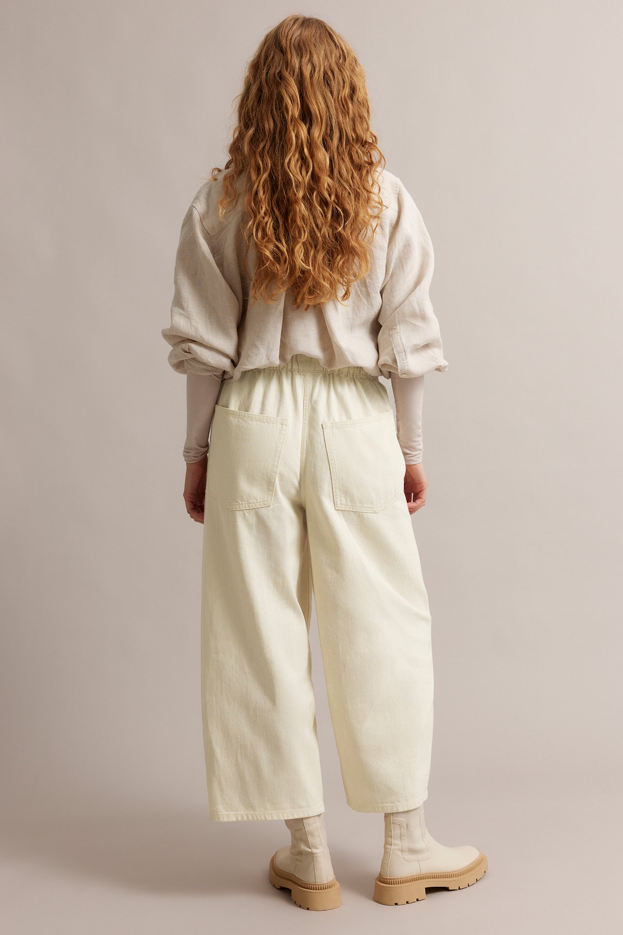 Widok z tyłu: kobieta w beżowych jeansach barrel leg z kieszeniami, jasnej bluzce i beżowych botkach.
