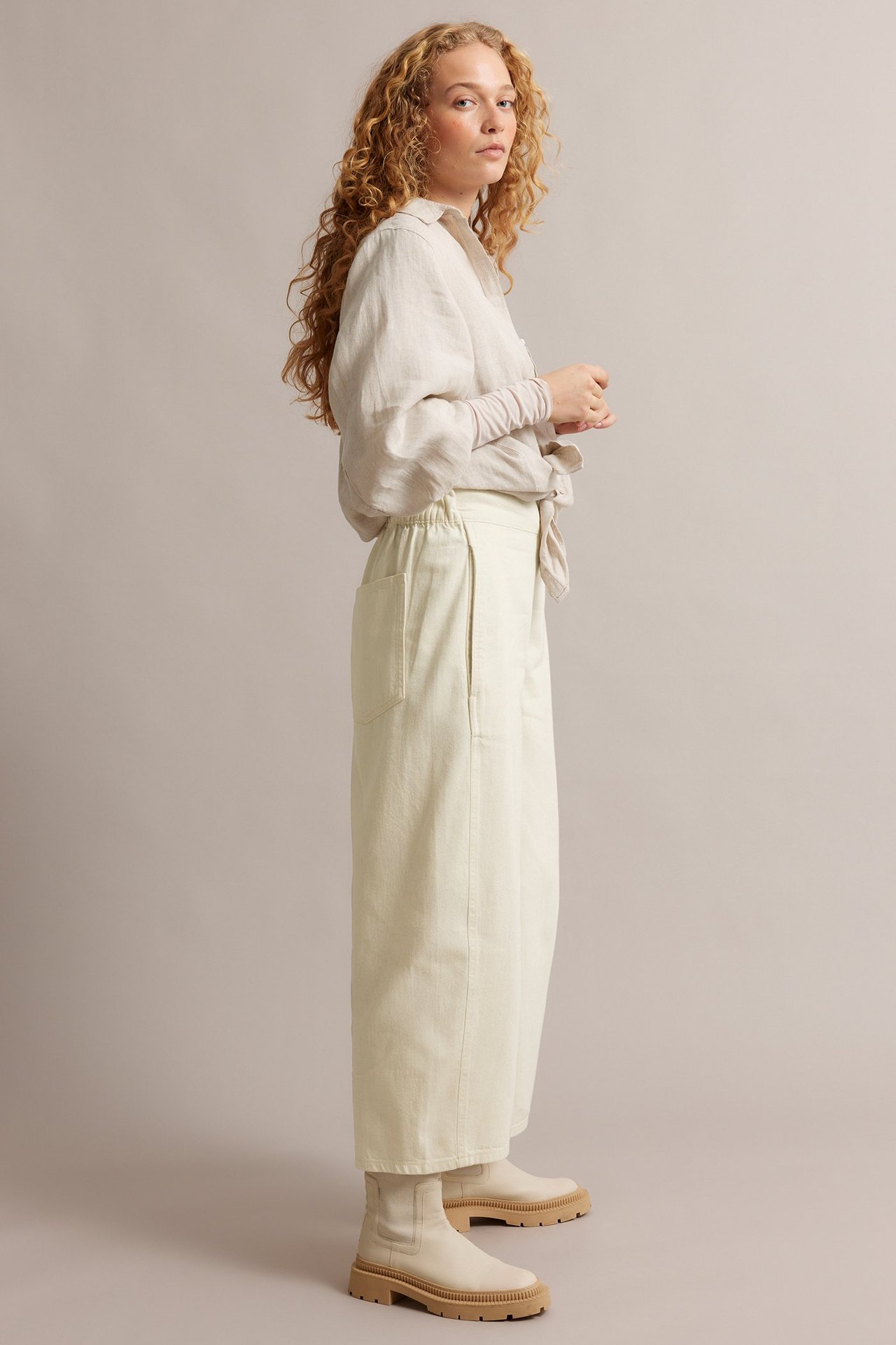 Nainen, sivukuva, beigenvärisissä barrel leg pull-on-farkuissa, vaaleassa paidassa ja beigenvärisissä saappaissa.