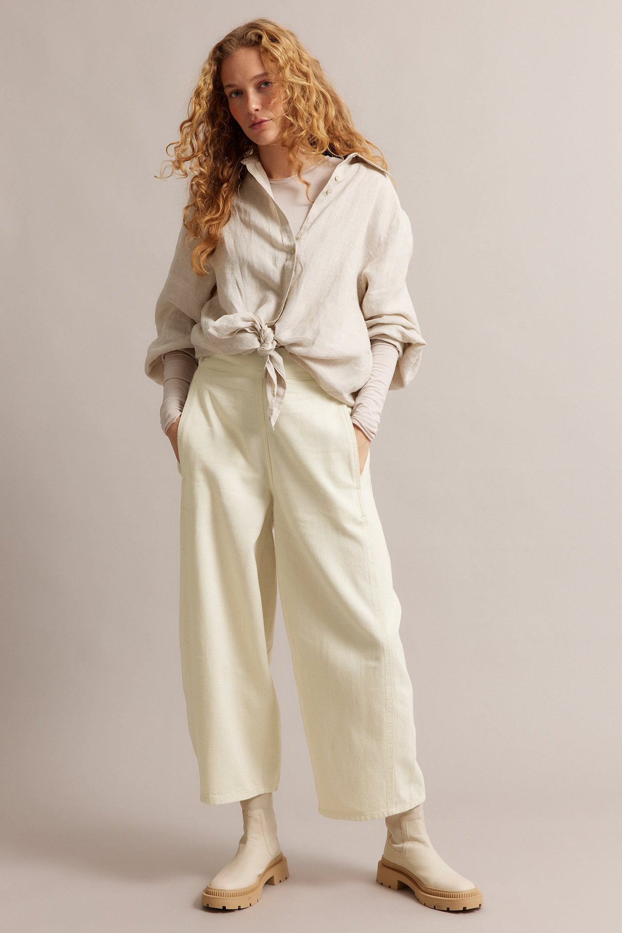 Widok z przodu kobiety w beżowych szerokich jeansach pull-on, beżowej koszuli wiązanej z przodu i beżowych botkach na platformie.