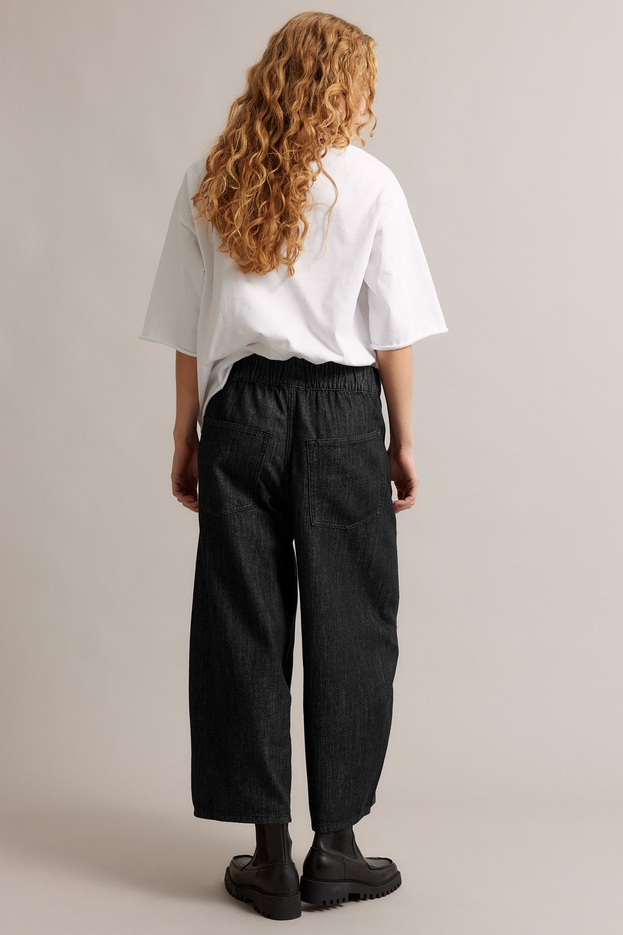 Ryggbilde av kvinne i svarte vide pull-on jeans i denim, hvit t-skjorte og svarte boots.