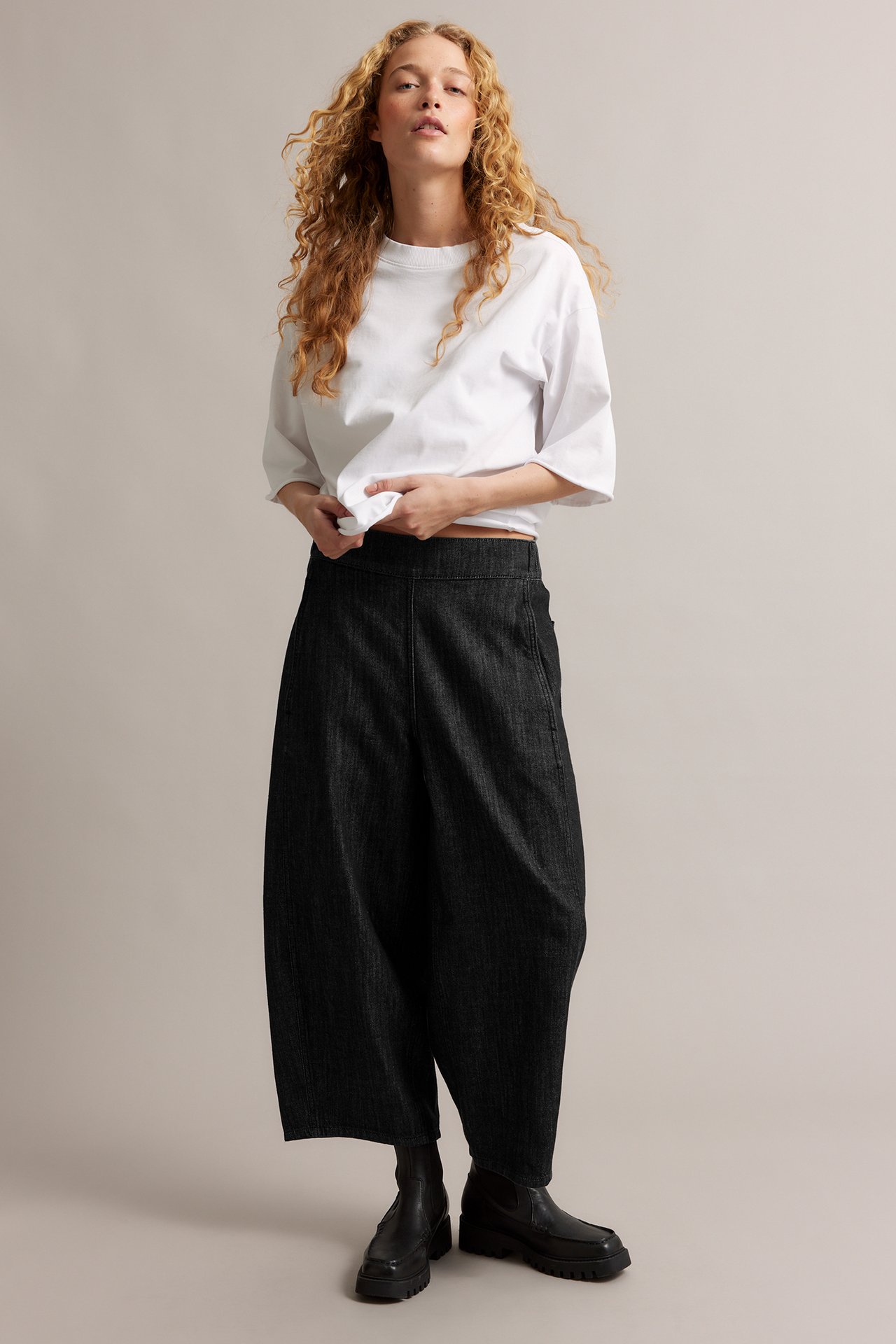 Frontvy av en kvinna i svarta barrel leg pull-on jeans i denim, vit t-shirt och svarta kängor.