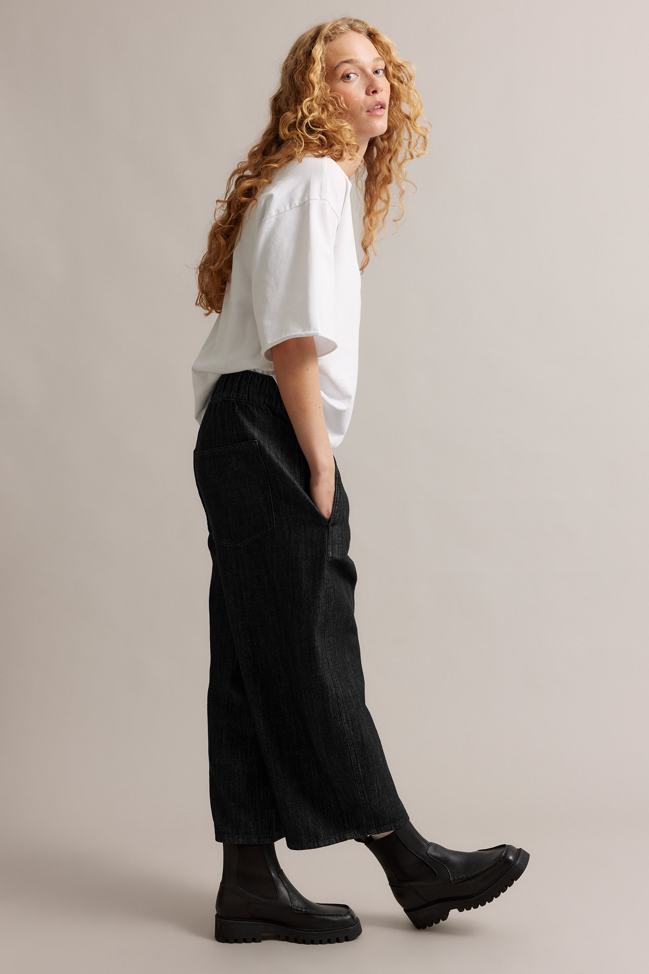 Kvinne i svarte barrel leg pull-on jeans, hvit t-skjorte og svarte chunky boots, sideprofil.