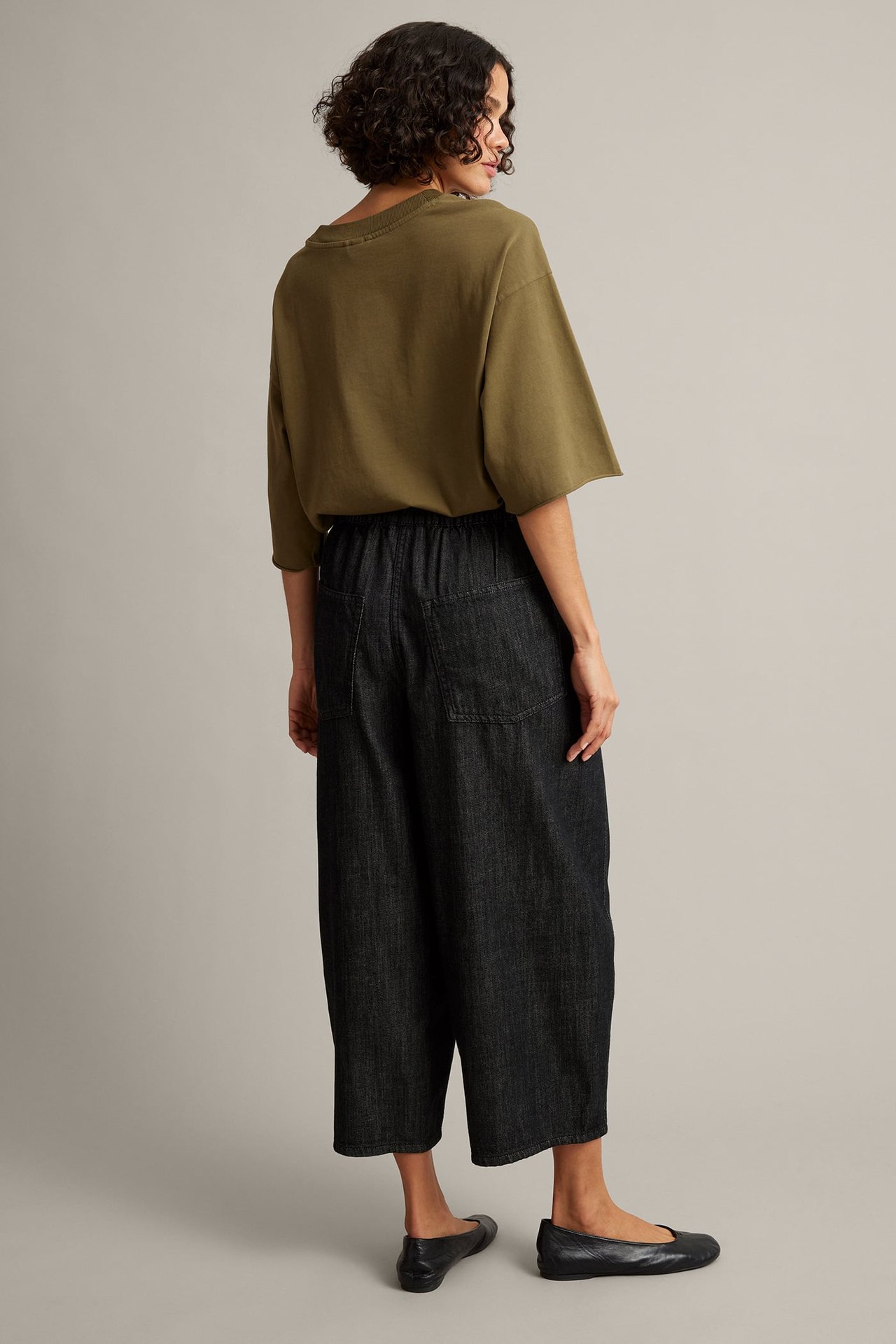 Bakifrån en kvinna i mörk tvätt vida beskurna byxor, olivgrön lös t-shirt och svarta ballerinaskor.