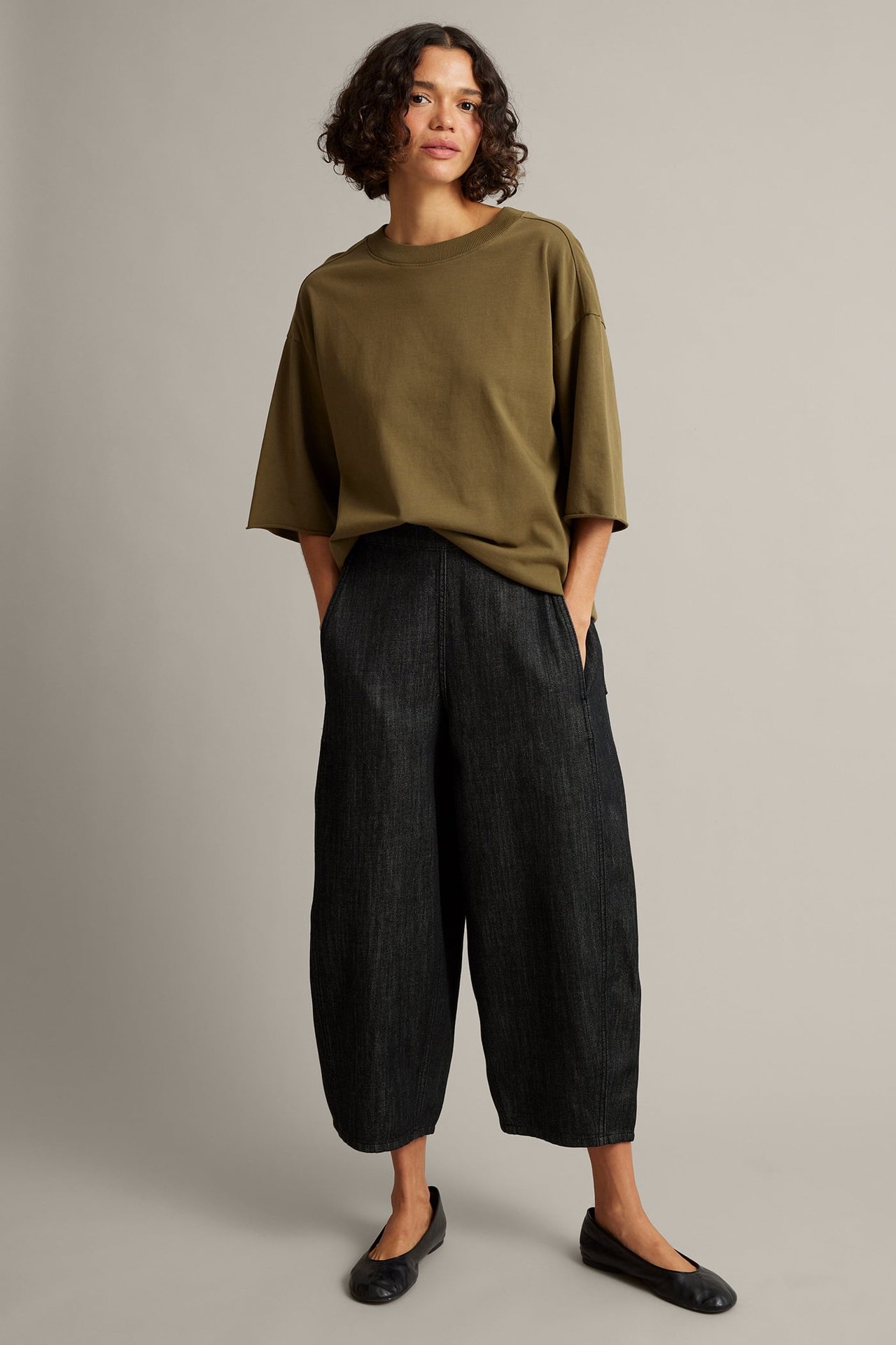 Frontvy av en kvinna i mörk tvätt vida beskurna byxor, en olivgrön oversize t-shirt och svarta platta skor.