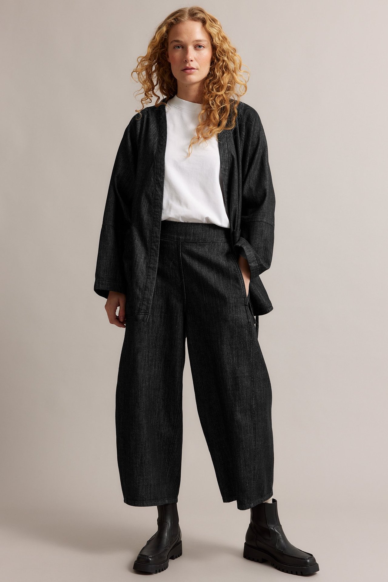Widok z przodu kobiety w czarnych jeansach barrel-leg, białym t-shircie, czarnej kurtce kimono i czarnych botkach.