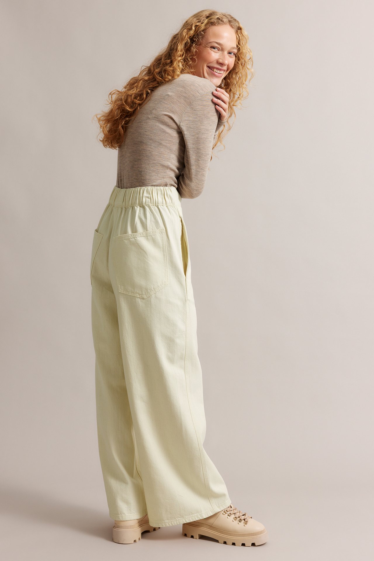 Widok z boku: kobieta w beżowych prostych dżinsach pull-on, jasnobrązowej bluzce i beżowych butach.