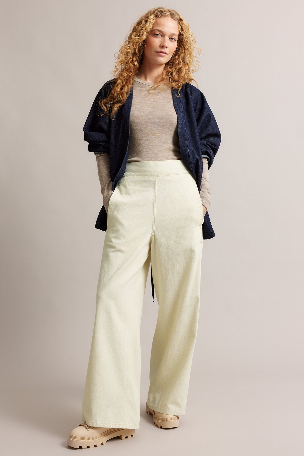 Frontvy: Kvinna bär ljusbeige raka pull-on-jeans, mörkblå jacka, ljusbeige topp, beigefärgade kängor.