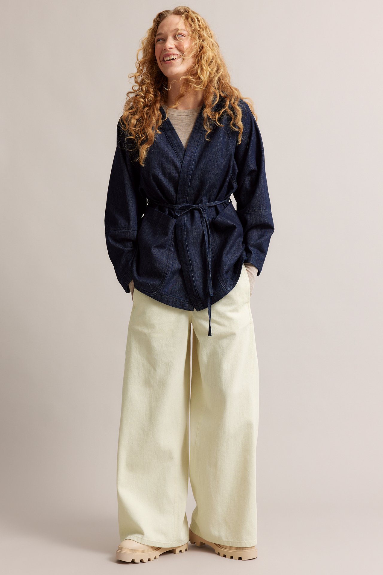 Framifrån, kvinna i beige vida pull-on jeans, mörkblå denimjacka med knytskärp och ljusbeige skor.