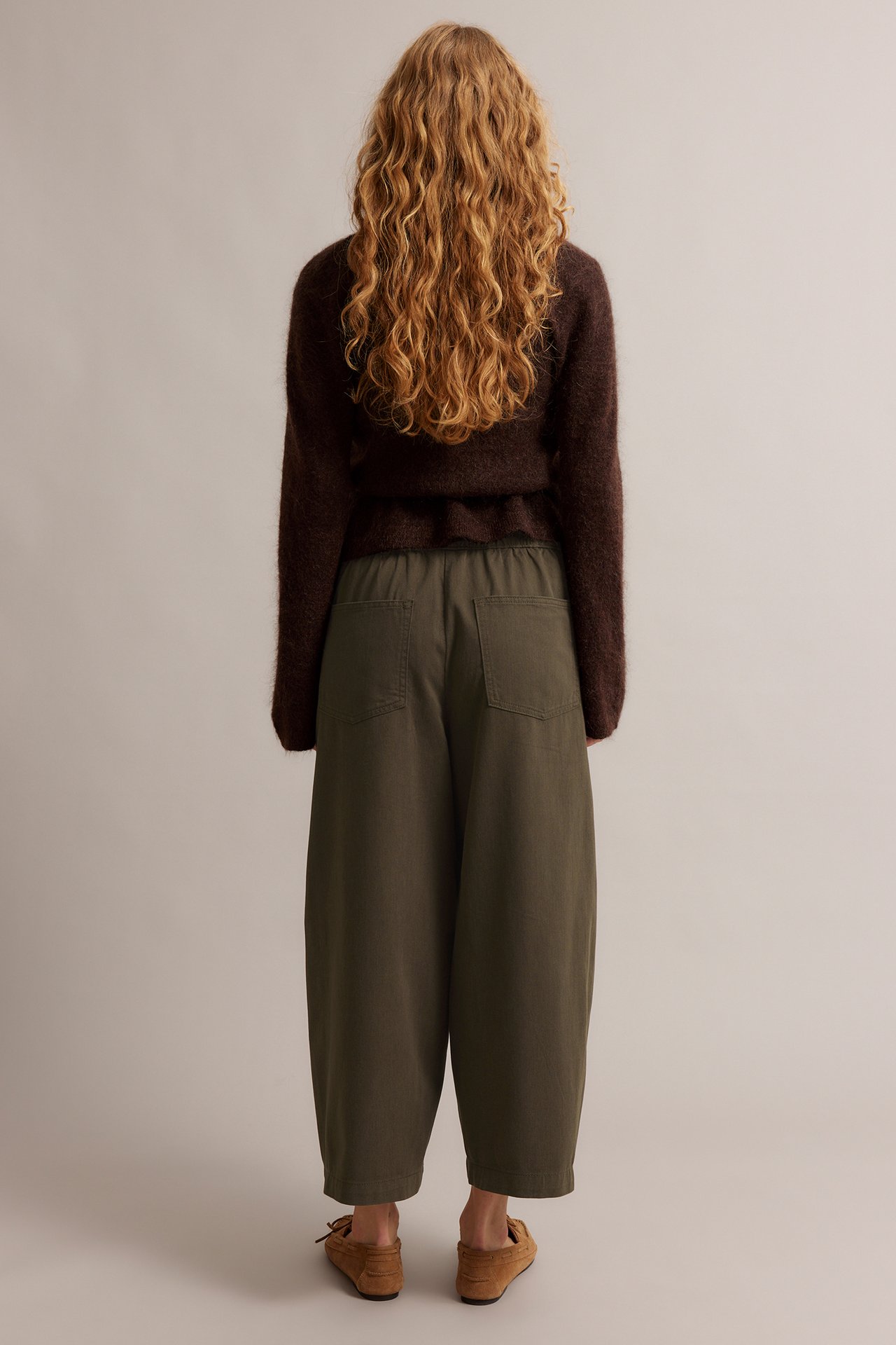 Widok z tyłu kobiety w ciemnooliwkowych spodniach pull-on, ciemnobrązowym swetrze i brązowych mokasynach.