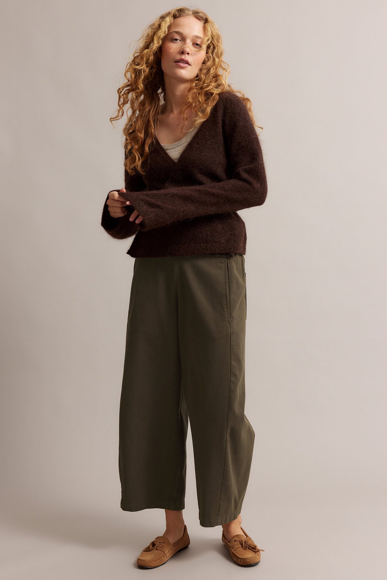 Edestä: nainen yllään oliivinvihreät leveälahkeiset pull-on-housut, ruskea pörröinen neuletakki ja beiget mokkasiinit.