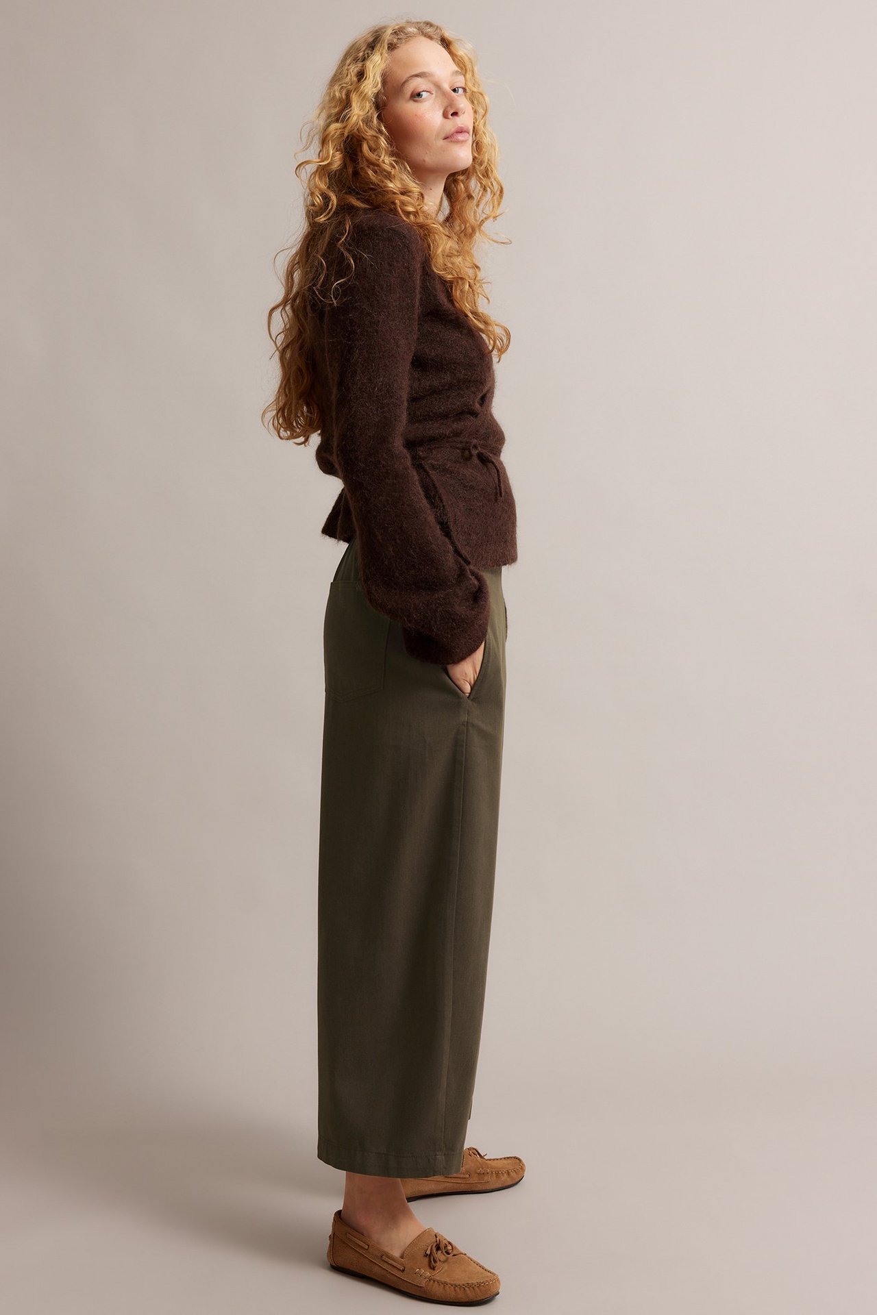 Naisen sivukuva yllään oliivinvihreät leveälahkeiset pull-on-housut, ruskea pörröinen neuletakki ja beiget mokkasiinit.