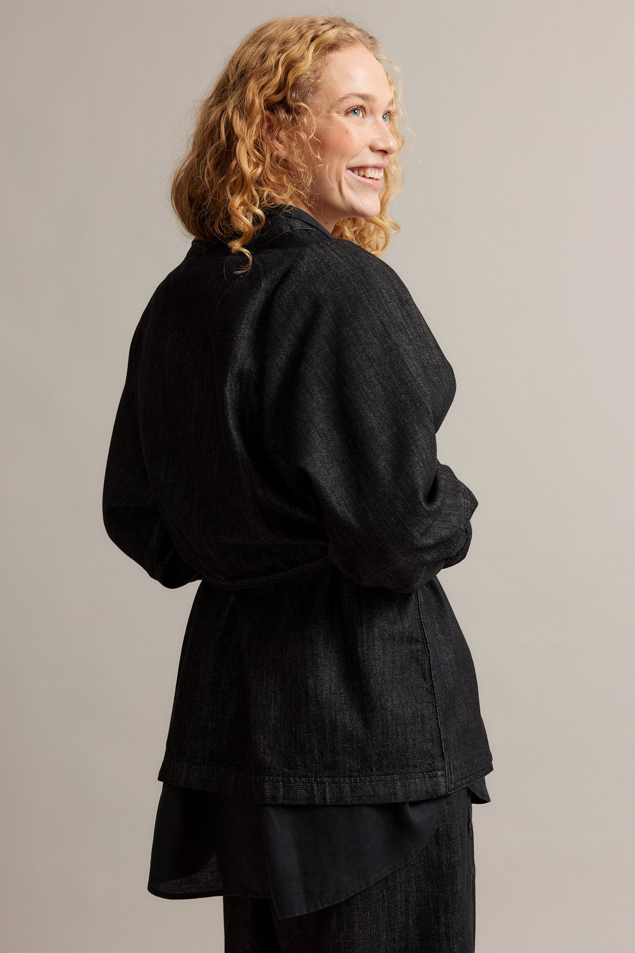 Kobieta z boku w czarnej denimowej kurtce kimono z wiązanym pasem, na ciemnym ubraniu.